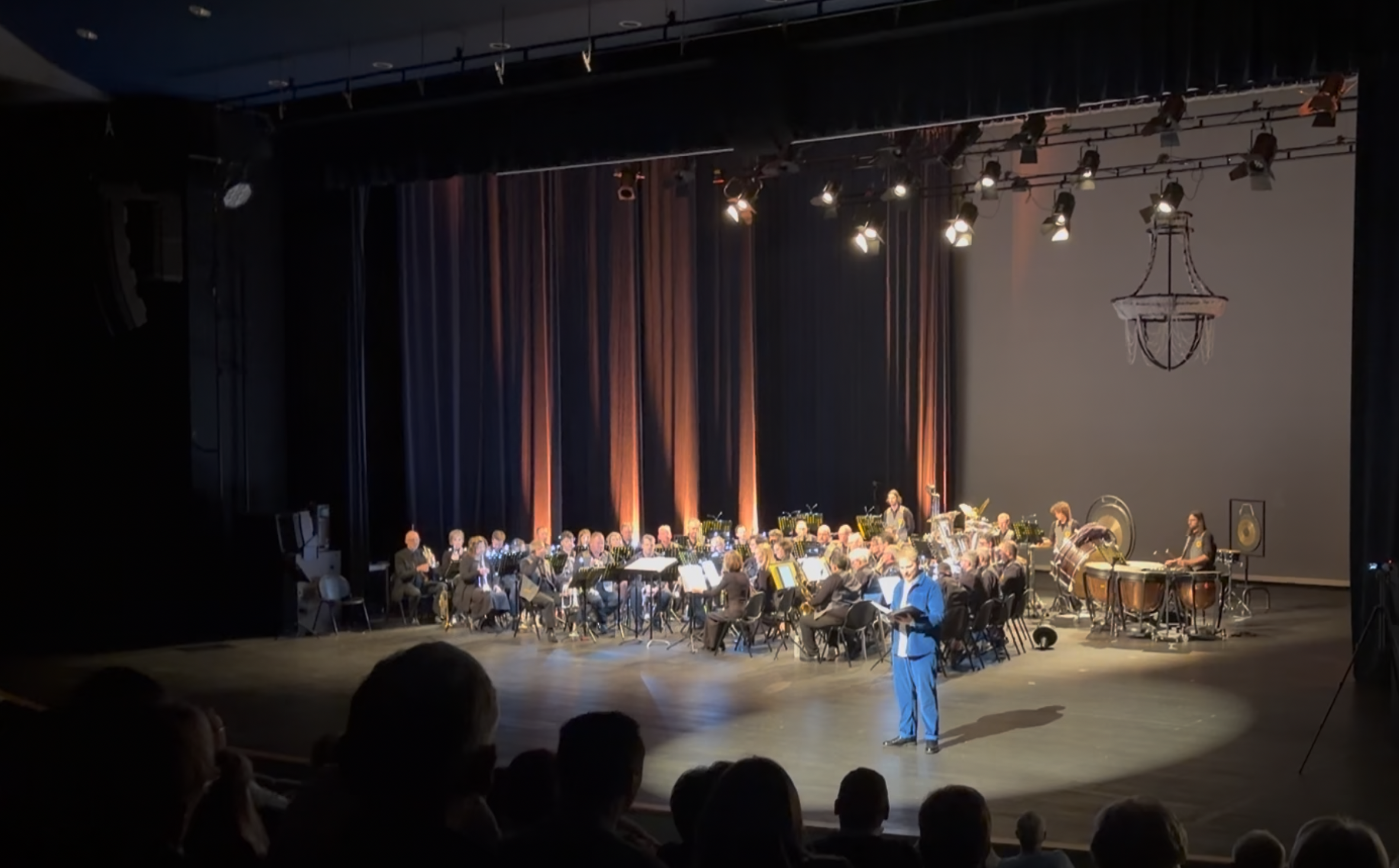 An orchestra performing on stage in a theater, with a conductor leading and stage lighting focused on the musicians. Audience members are visible in the foreground.