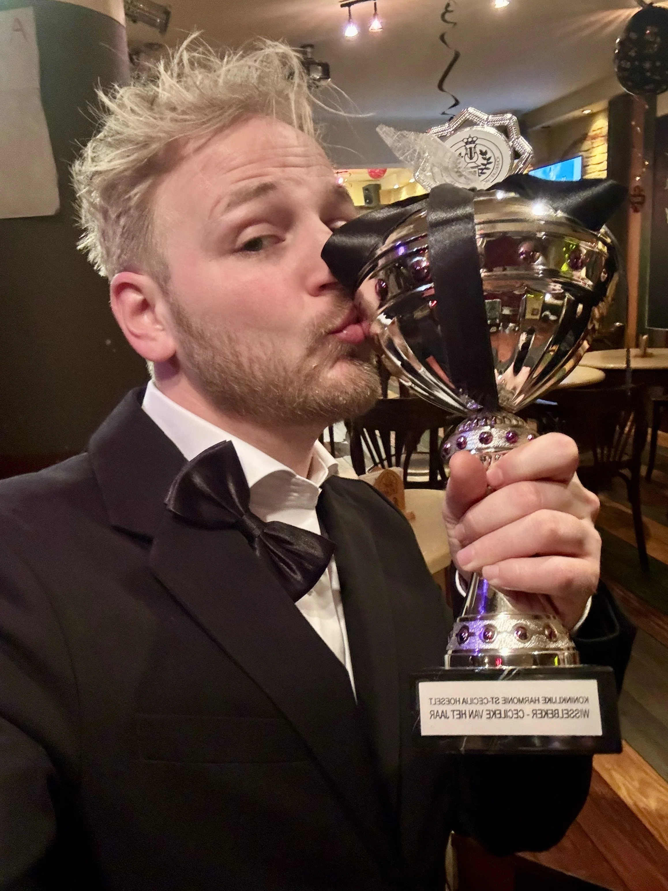 A man in a tuxedo is holding and kissing a trophy with a reflective silver surface, black accents, and a decorative base with purple jewels. The trophy has markings and text on it, but they are not fully visible in the image.
