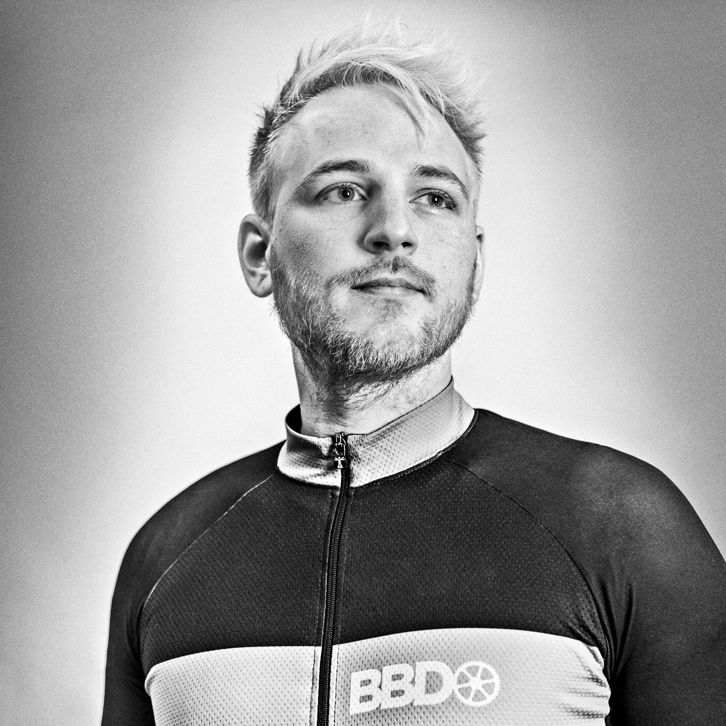 Close-up of a male cyclist's face and upper body, wearing a black and white cycling jersey with the 'BBD' logo, against a neutral background.