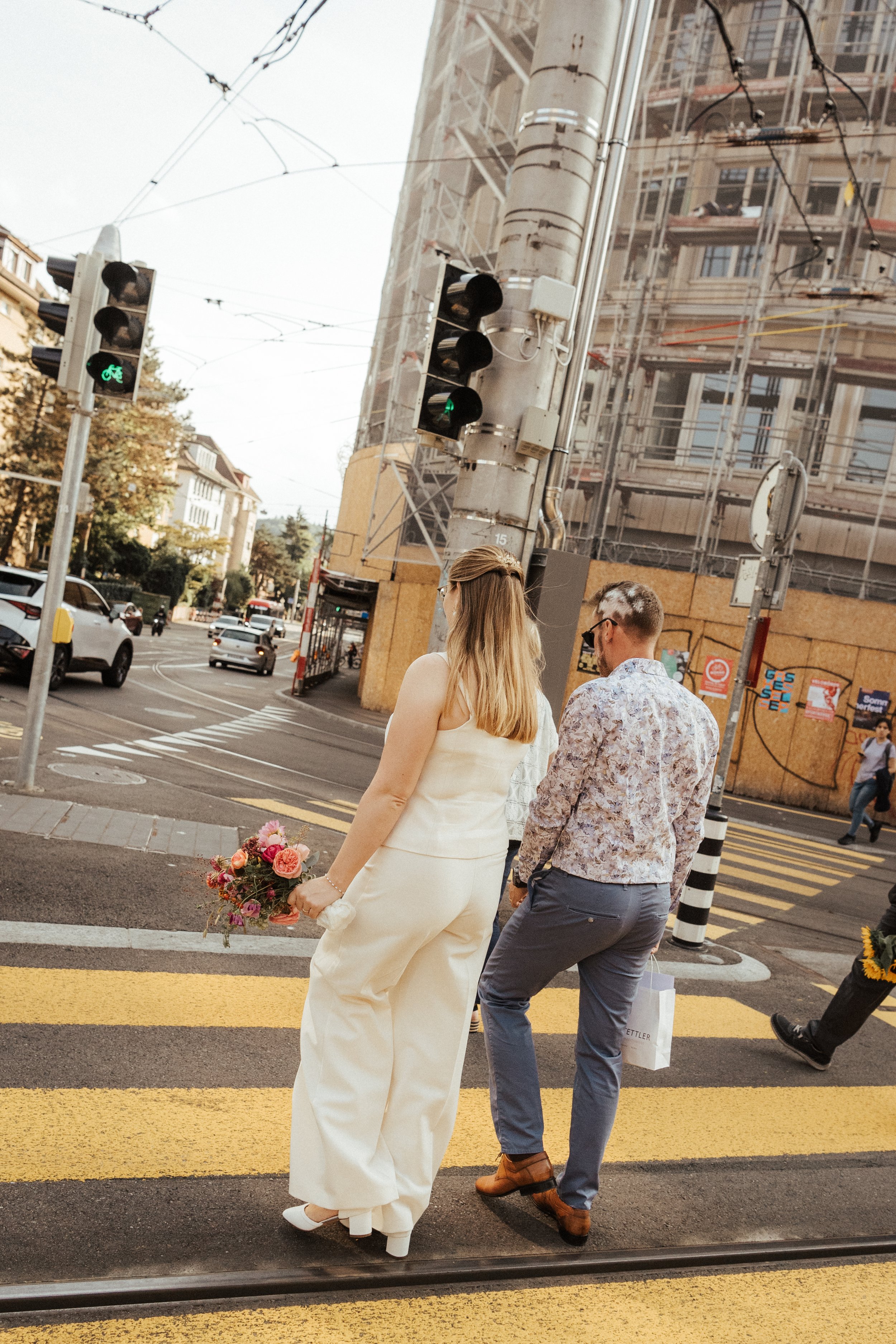 Ein Brautpaar überquert eine Straße an einer Ampel bei Rotlicht. Die Braut trägt ein cremefarbenes Hochzeitskleid und hält einen Blumenstrauß. Der Bräutigam trägt ein florales Hemd und dunkle Hose. Es ist Tag, und im Hintergrund sind Autos, eine Baus
