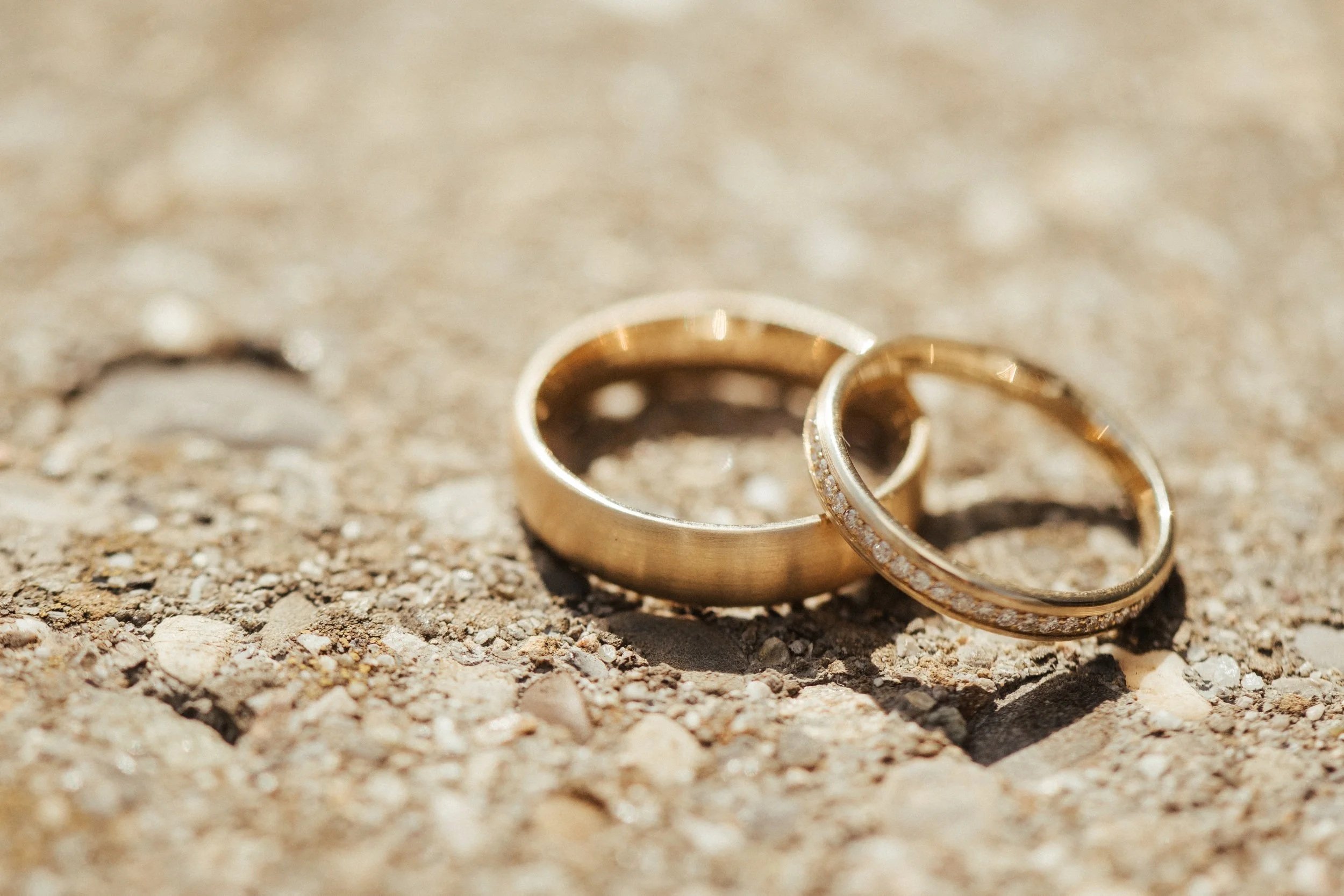Zwei goldene Eheringe liegen auf einem Sandboden, einer ist mit kleinen Diamanten besetzt.