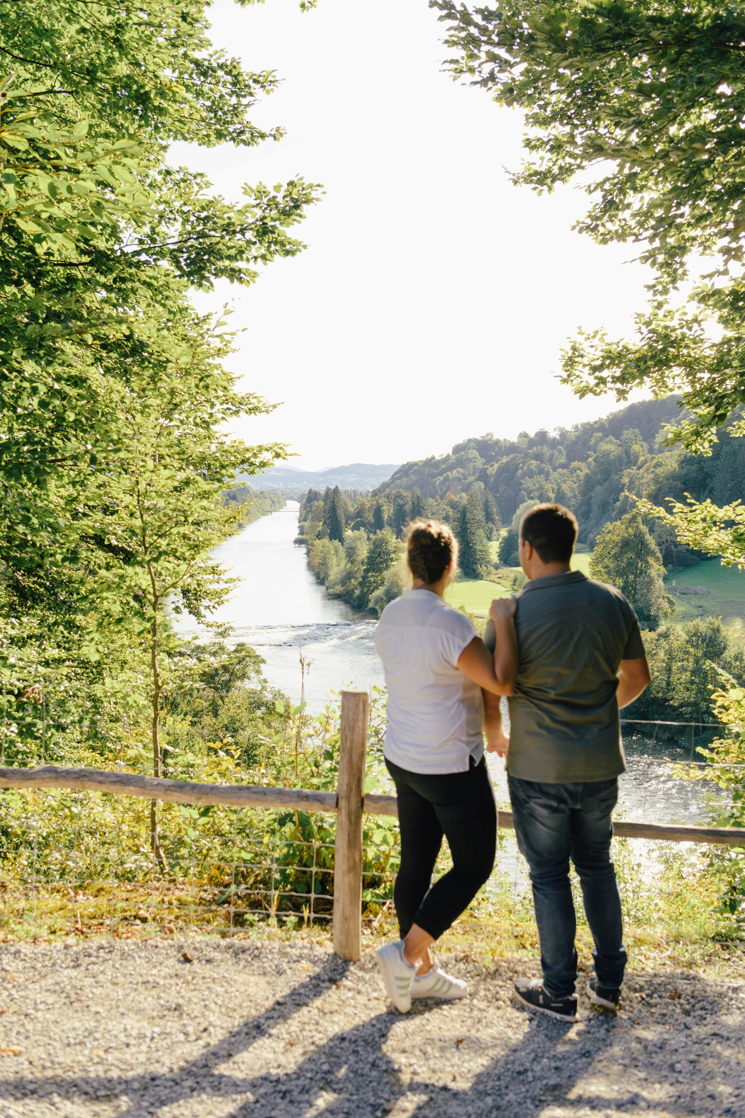 Zwei junge Männer stehen an einem Wasserfall mit Blick auf einen Fluss, umgeben von grünen Bäumen, bei Sonnenschein, während sie sich an einem Holzzaun lehnen.
