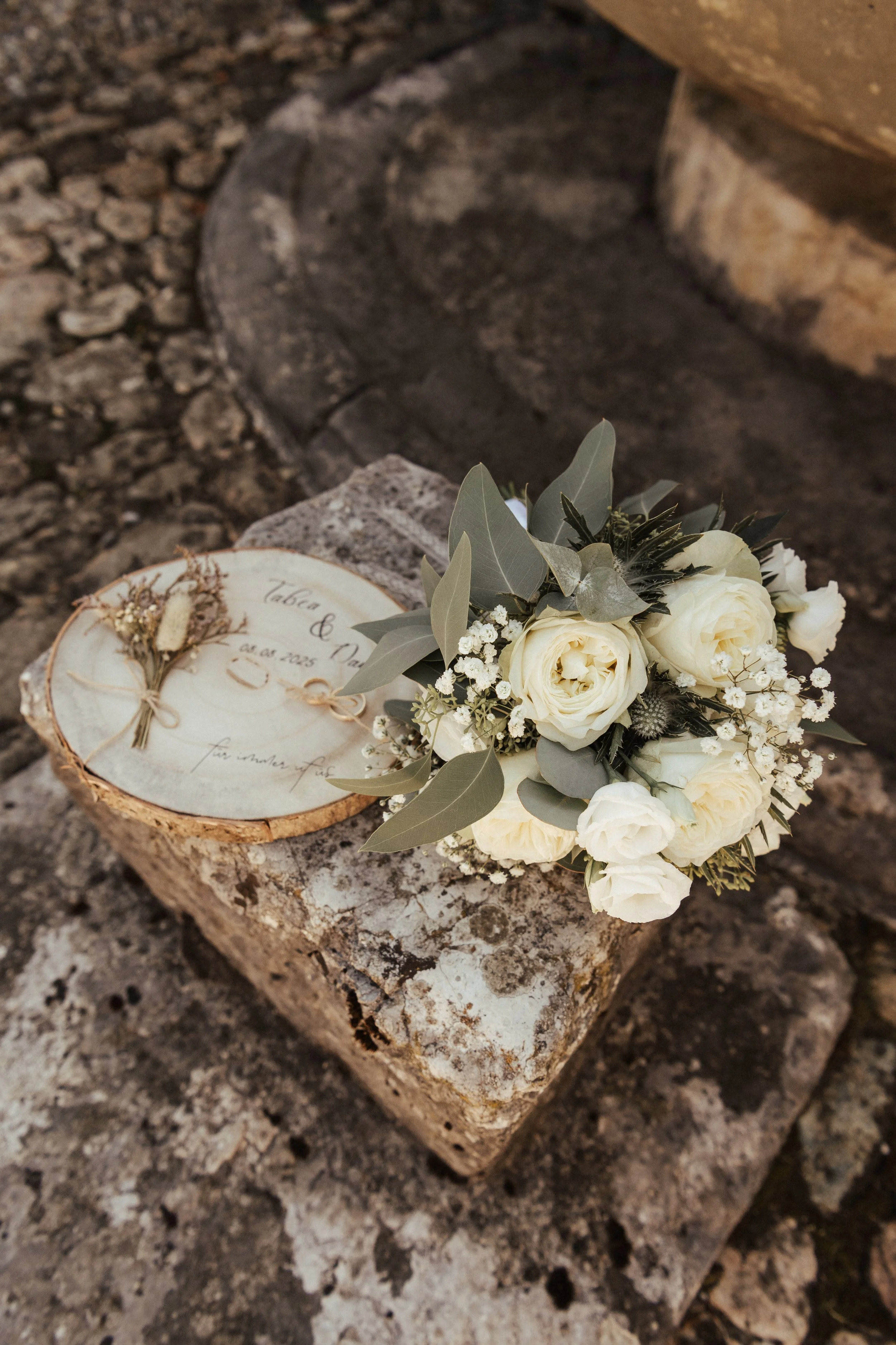 Handgefertigte Hochzeitseinladung auf einem Holzscheibchen, verziert mit weißen Rosen, Eukalyptus und anderen Blumen, liegt auf einem Stein.