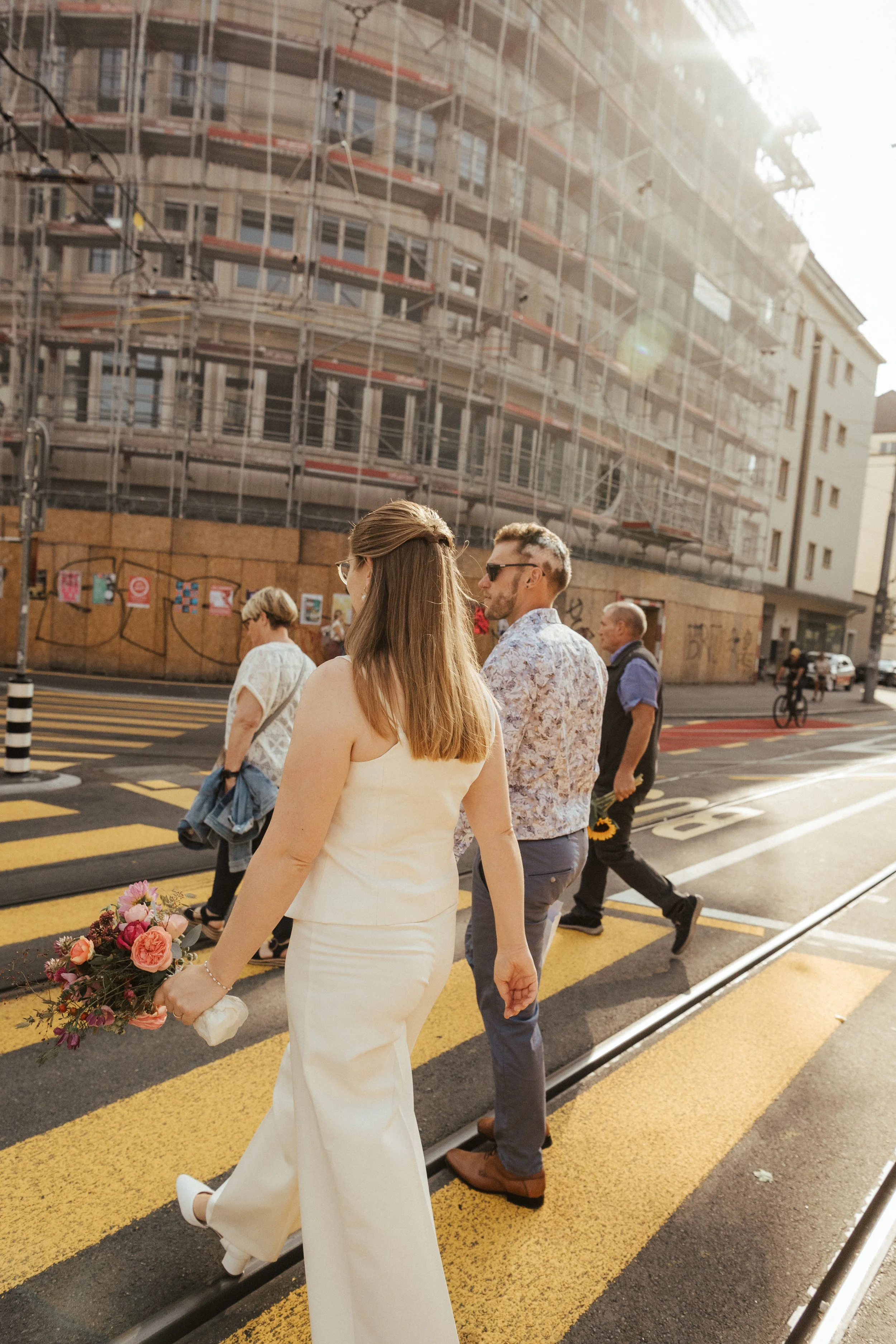 Eine Frau in einem weißen Kleid hält einen Blumenstrauß. Sie geht an einer Gruppe von Leuten vorbei, die auf einer Kreuzung auf die Straße treten. Im Hintergrund ist ein Gebäude mit Gerüst und Graffiti auf einer Holzwand.