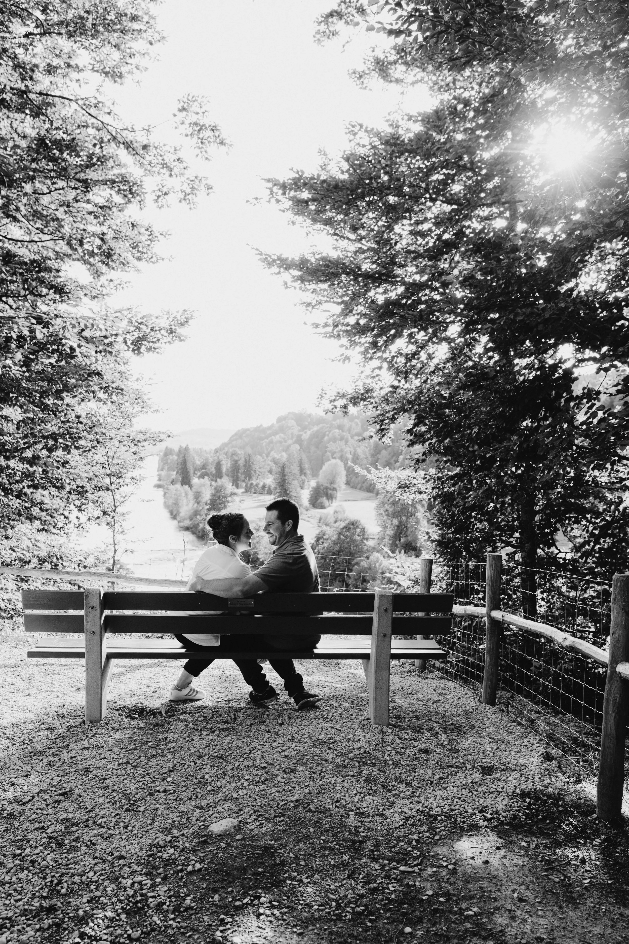 Ein Mann und eine Frau sitzen auf einer Parkbank im Freien, umgeben von Bäumen und einer Landschaft mit Hügeln im Hintergrund, während die Sonne durch die Blätter scheint.