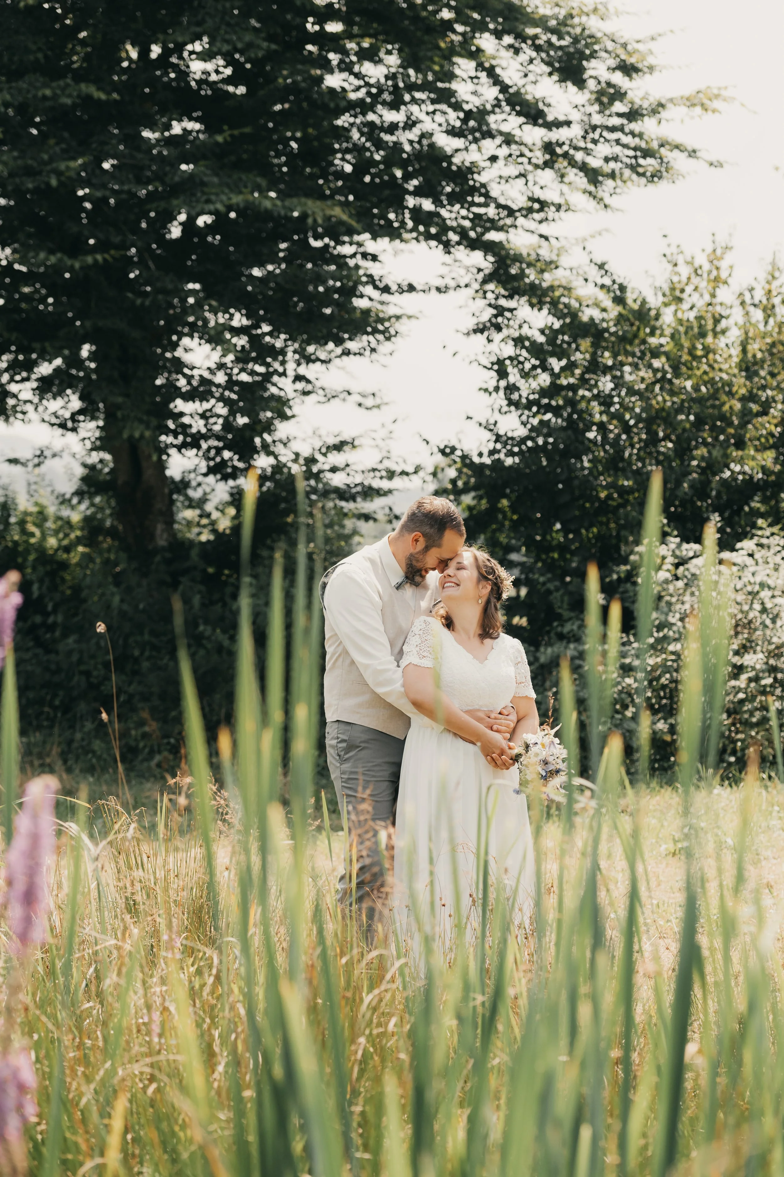 Ein glückliches Paar bei einer Hochzeit im Freien, umgeben von grünem Gras und Bäumen, die sich umarmen und lachen.