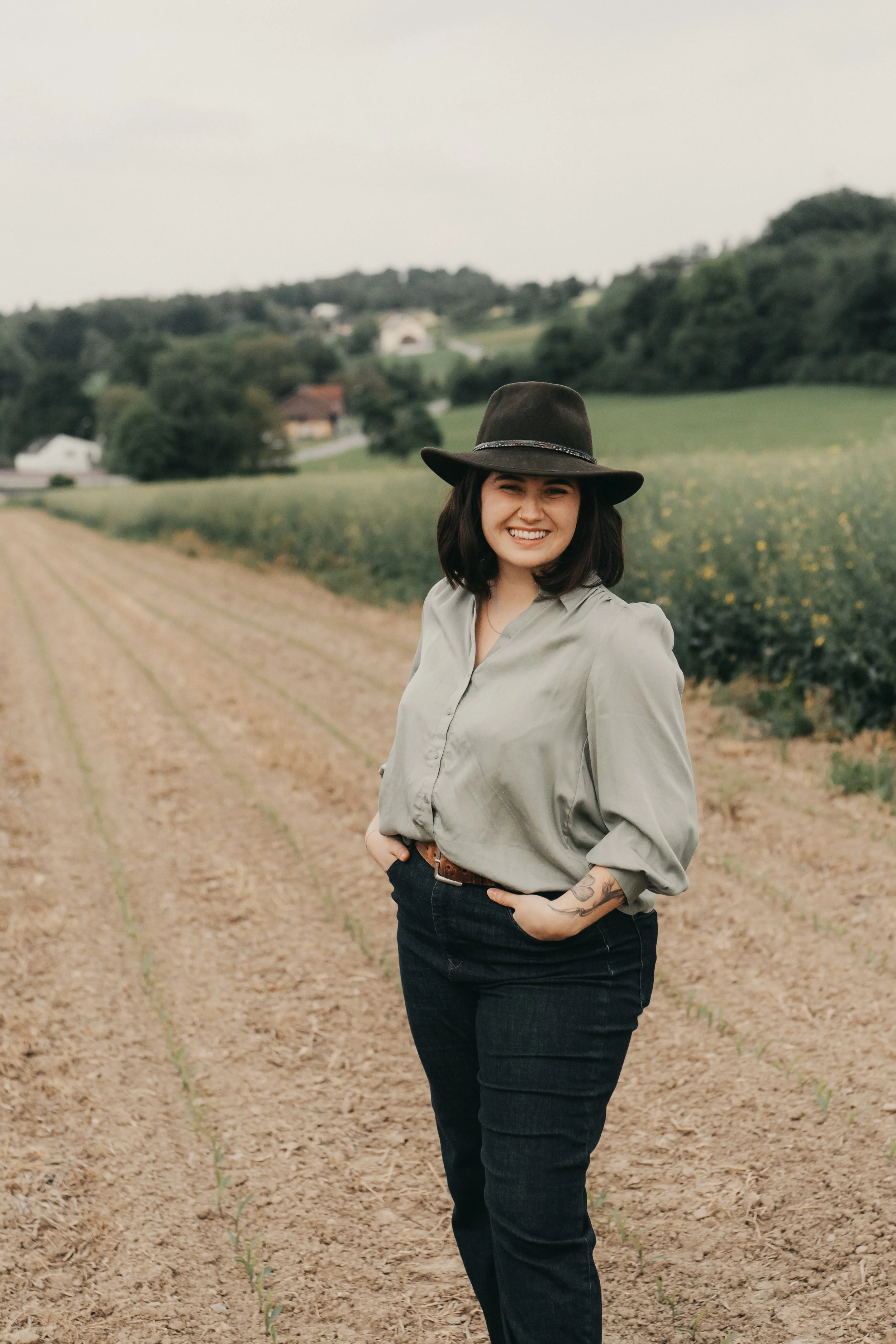 Lächelnde Frau mit dunklem Hut und Tattoos, stehend in einem Feld mit jungen Pflanzen, im Hintergrund Hügel und Häuser.