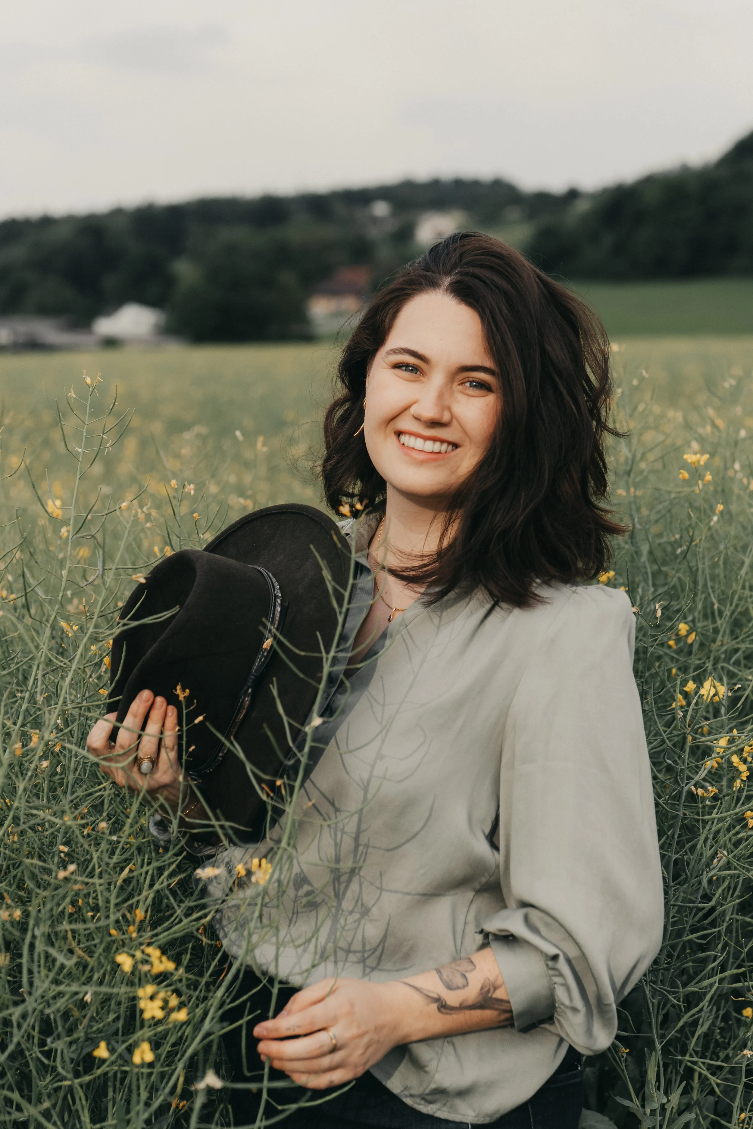 Junge Frau mit dunklen Locken und Tattoos im Feld mit gelben Blumen, lächelnd, trägt hellgraue Bluse und hält schwarzen Hut.