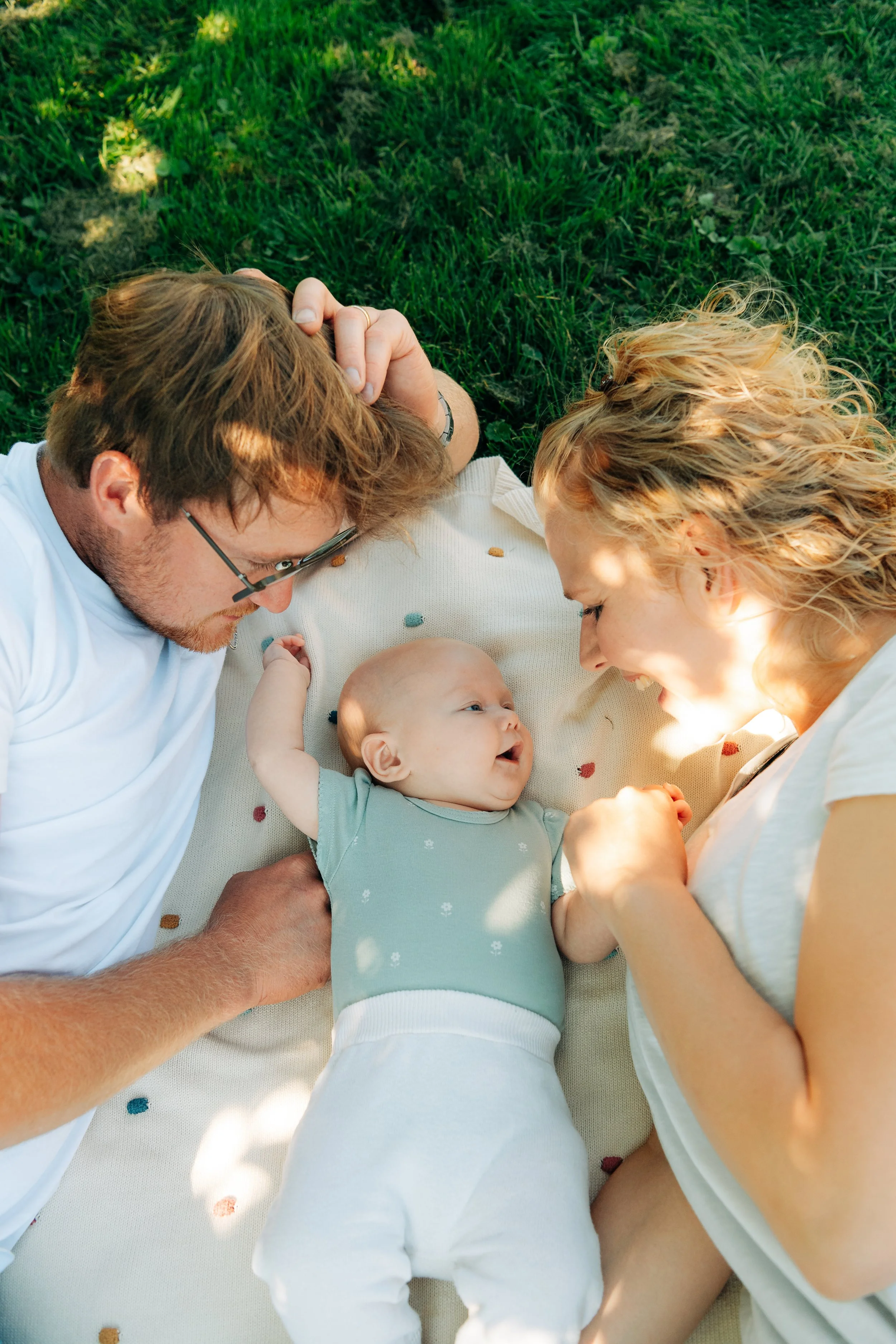 Familie liegt an einem sonnigen Tag auf einer grünen Wiese und schaut fröhlich auf ein neugeborenes Baby.