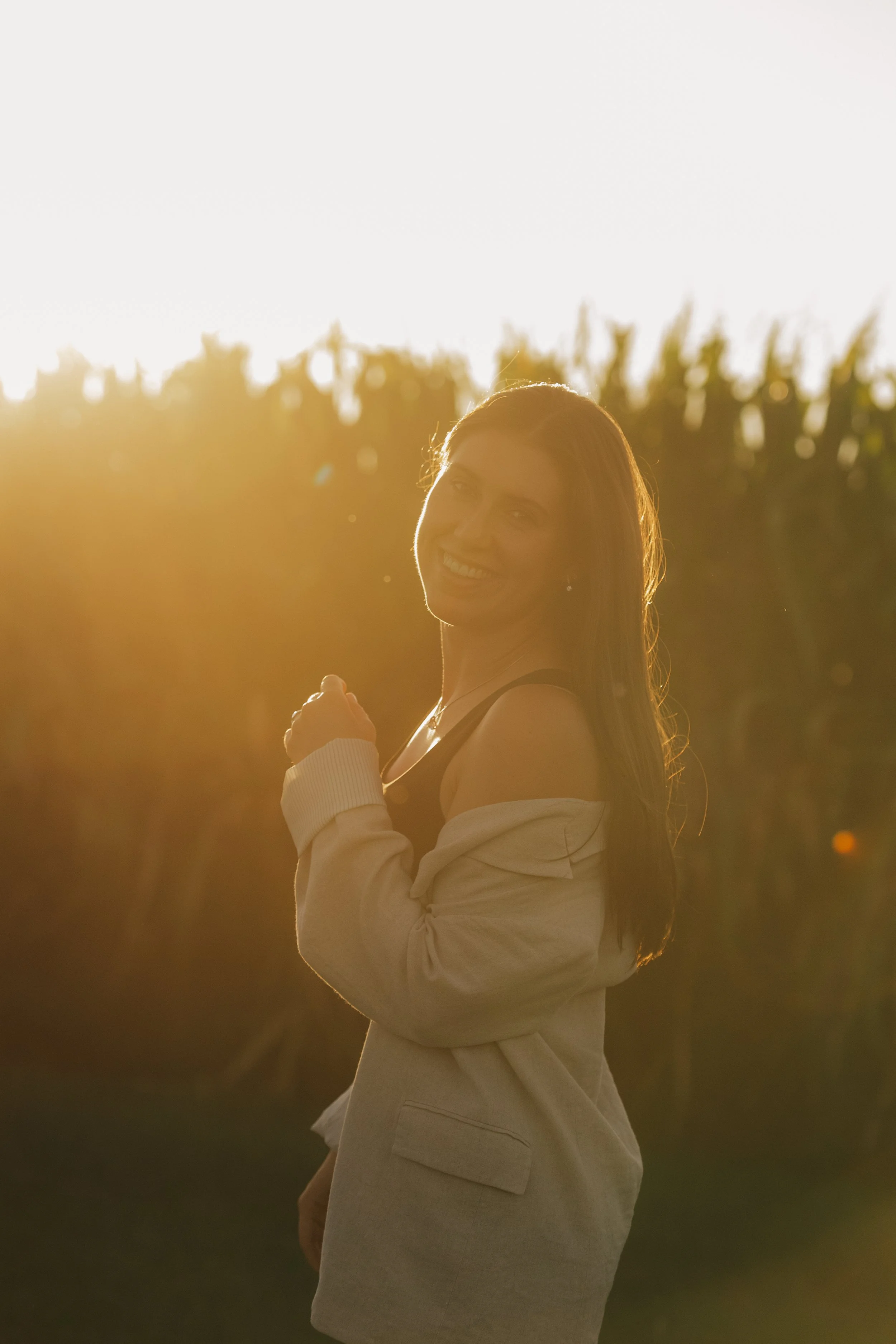 Eine lachende Frau mit langen braunen Haaren steht im Sonnenlicht in einem Feld mit Bäumen im Hintergrund.