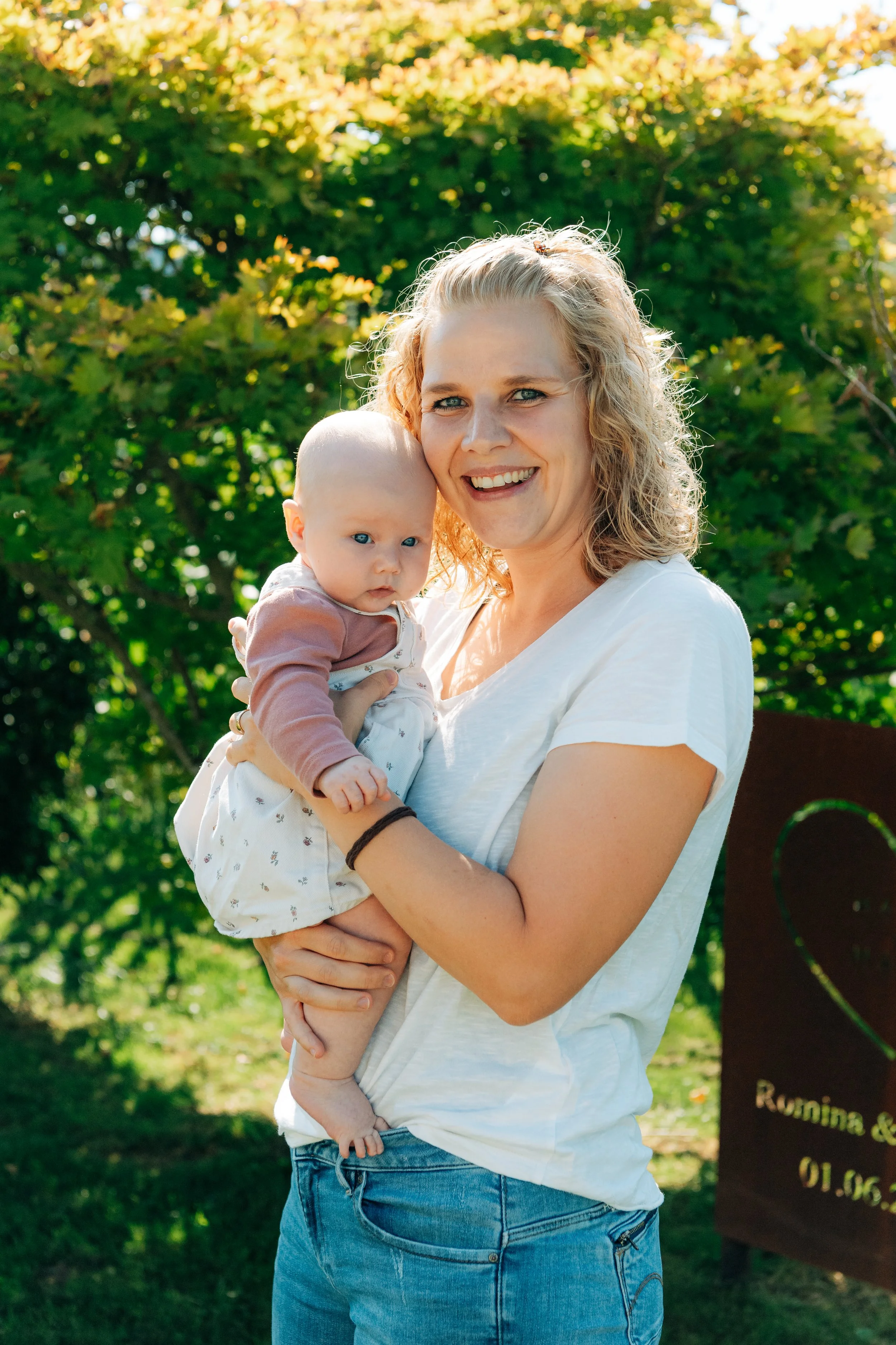 Eine lachende Frau mit blonden, gewellten Haaren hält ein Baby mit roten Haaren und blauen Augen in ihren Armen. Sie sind im Freien, im Sonnenschein, vor grünen Bäumen.