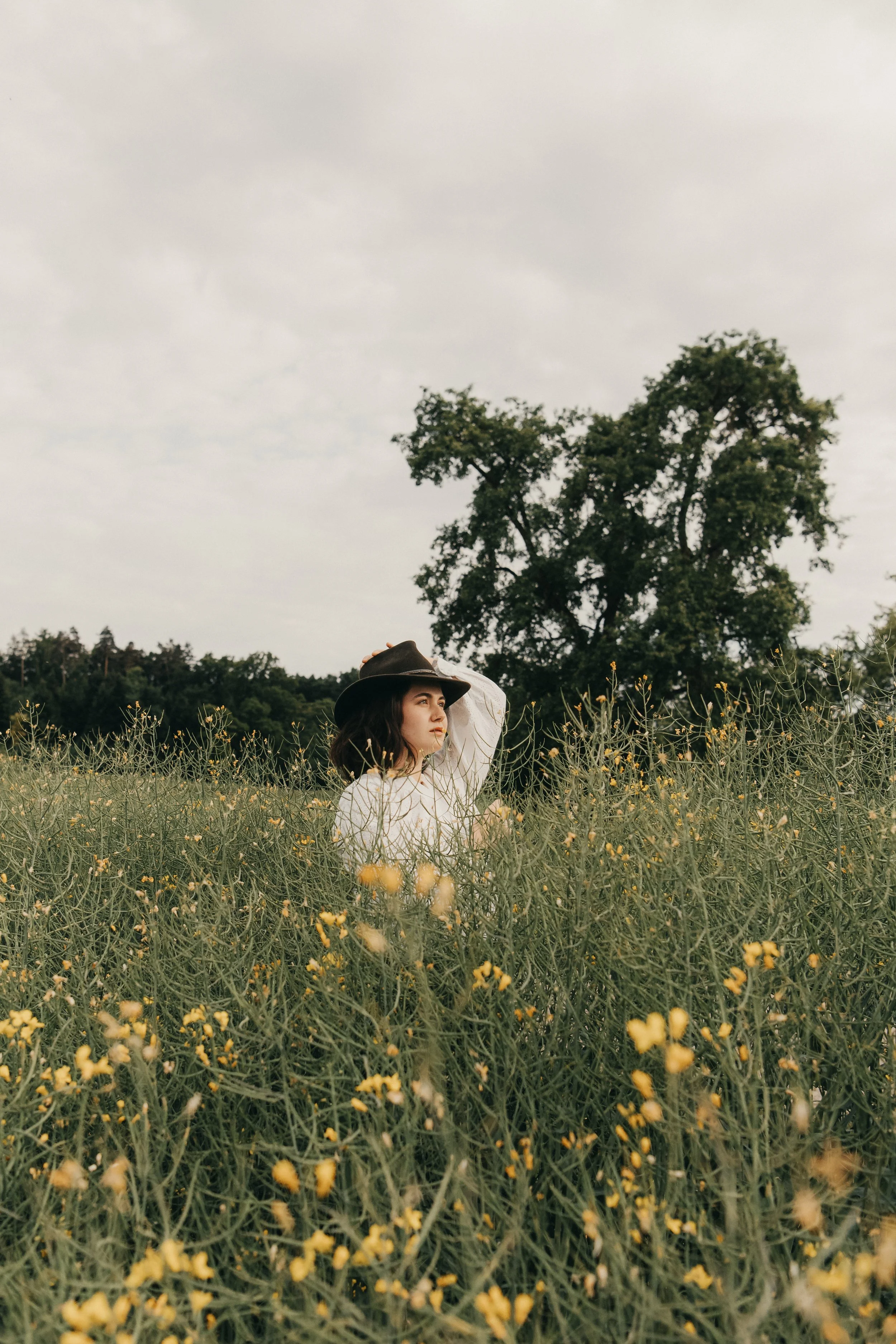 Eine junge Frau mit braunen Haaren trägt einen Strohhut und steht in einem Feld mit kleinen gelben Blumen, im Hintergrund sind große Bäume und ein grauer Himmel.