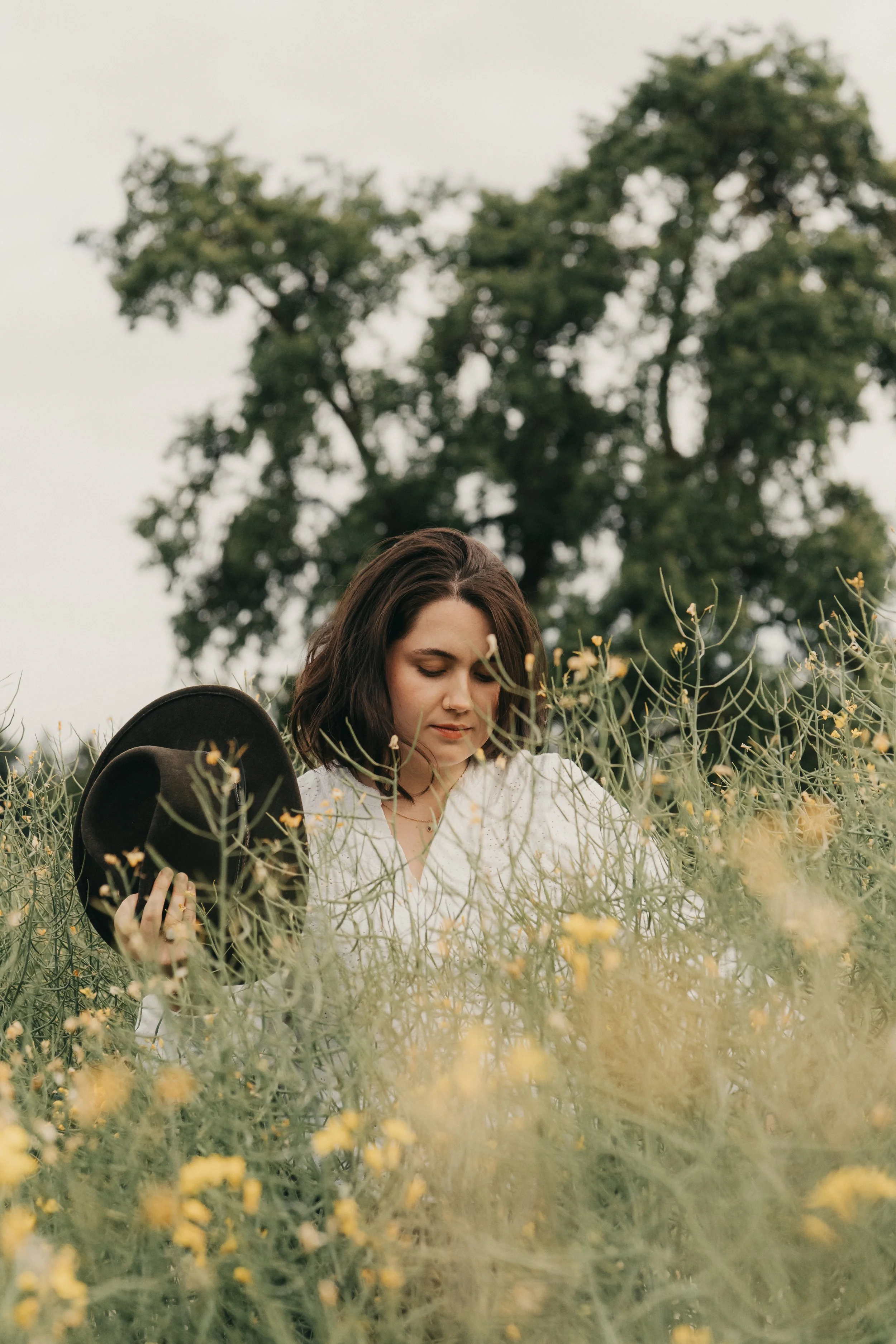Ein junges Mädchen mit braunen Haaren steht in einem Feld mit gelben Blumen, hält einen schwarzen Hut in der Hand und sieht nach unten, im Hintergrund ein großer Baum und ein grauer Himmel.