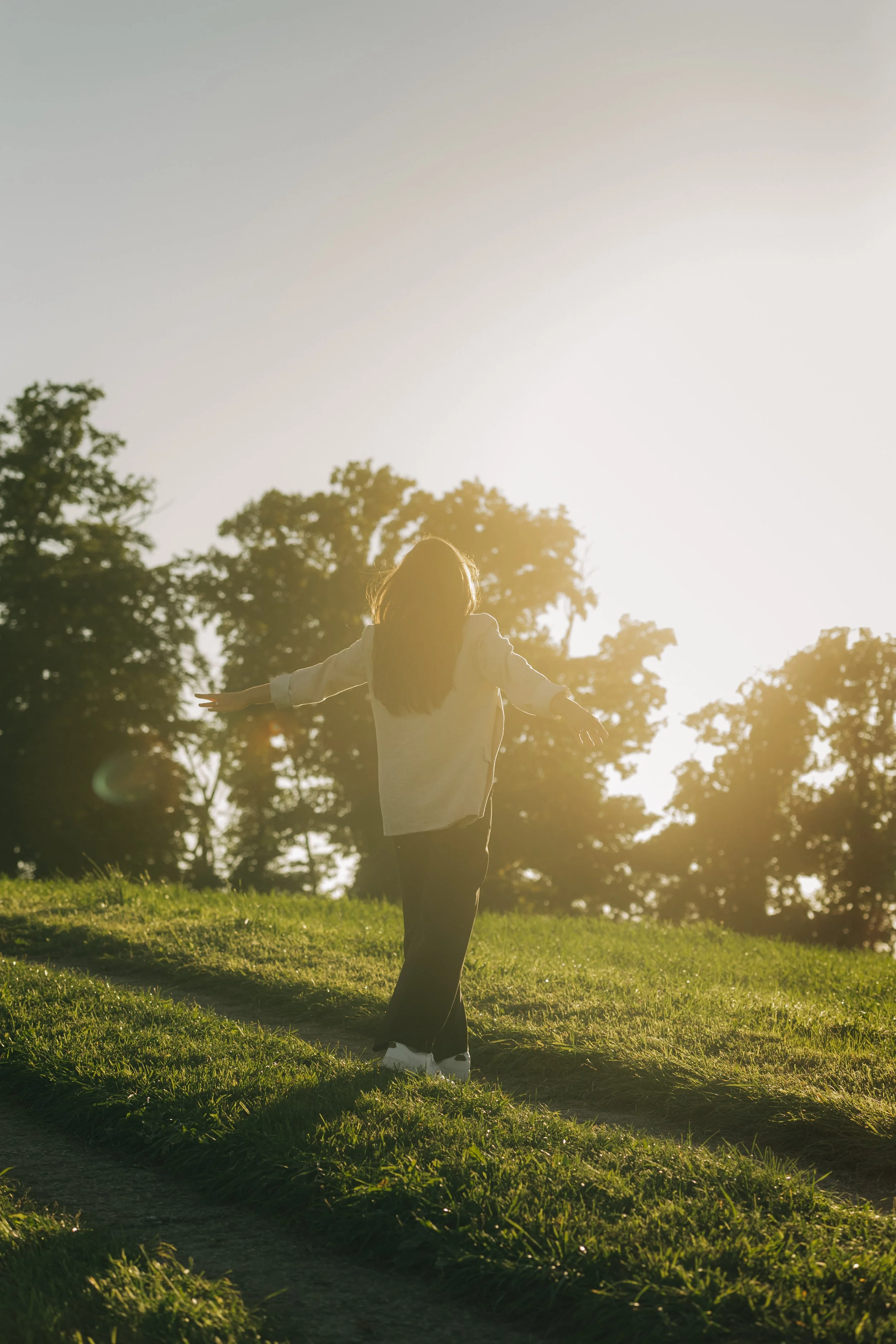 Eine Person läuft im Sonnenlicht durch ein grünes, offenes Feld mit Bäumen im Hintergrund. Die Person trägt eine dunkle Hose und ein helles Oberteil, die Arme sind ausgebreitet.