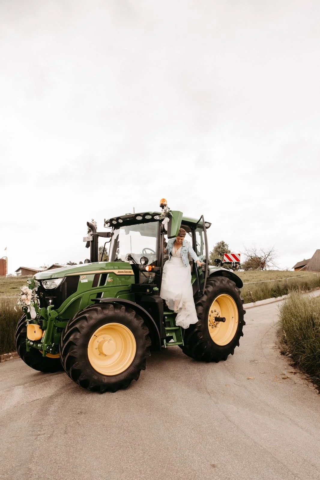 Eine Frau in einem weißen Brautkleid steigt aus einem grünen Traktor, der mit Dekorationen geschmückt ist, auf einer ländlichen Straße.