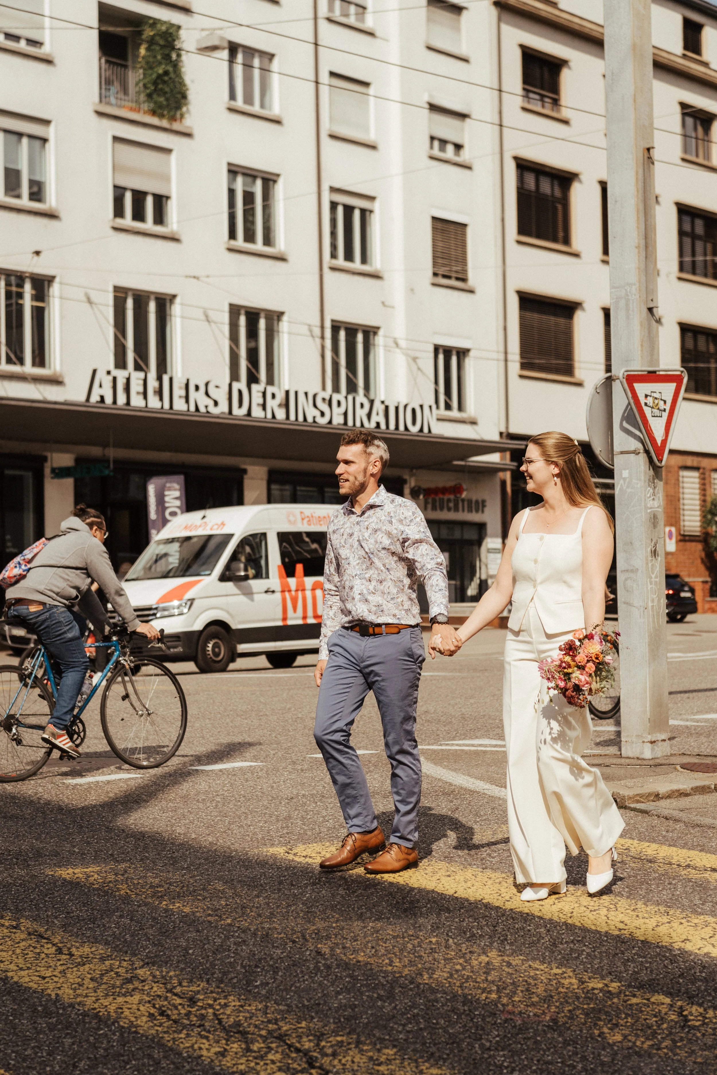 Ein frisch vermähltes Paar hält Händchen beim Überqueren einer Straße in der Stadt, umgeben von Passanten, Fahrradfahrern und einem Lieferwagen.