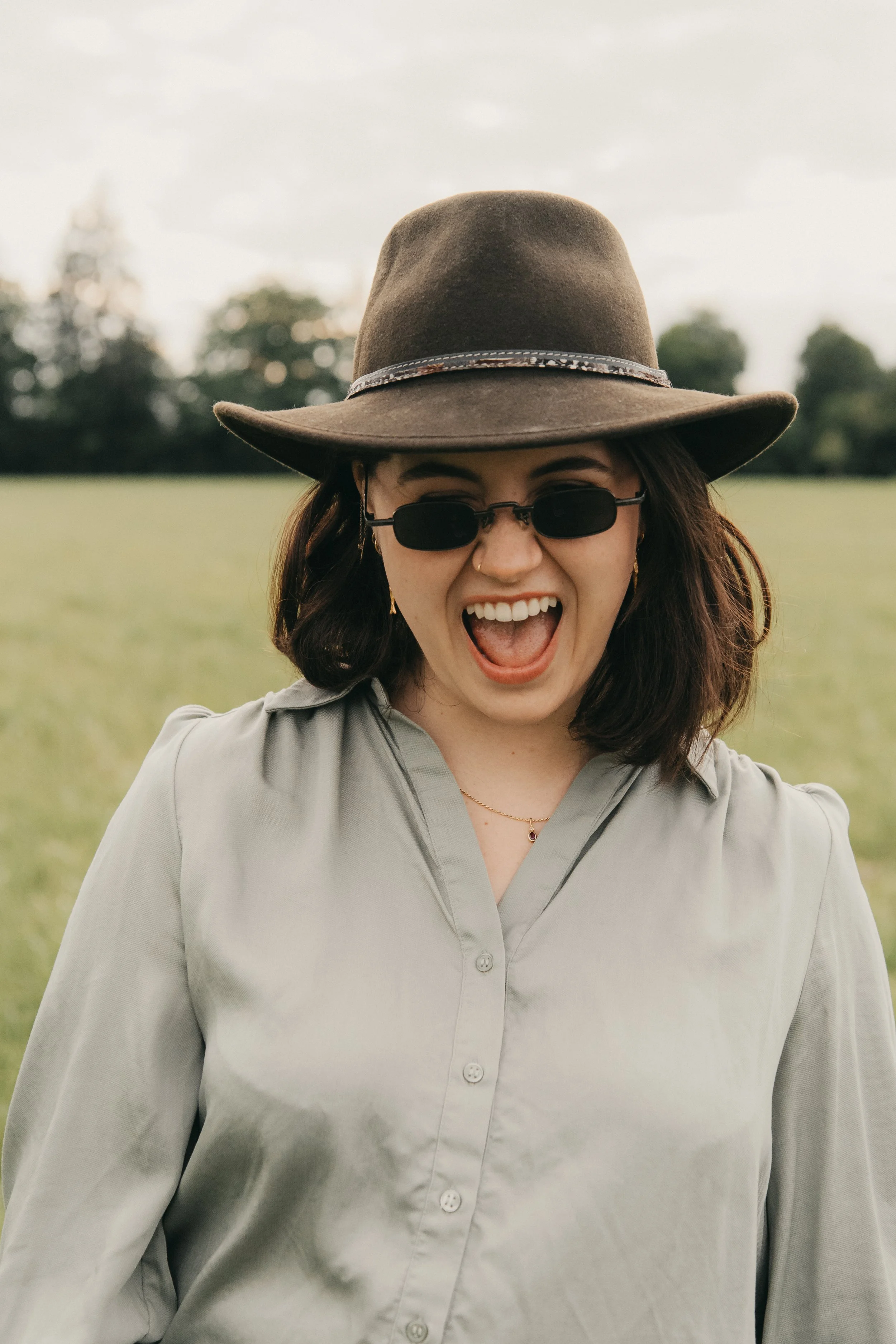 Junge Frau mit braunen Haaren, schwarzer Sonnenbrille, grauem Hemd und Hut, lachend im Freien, im Hintergrund Bäume und Himmel.
