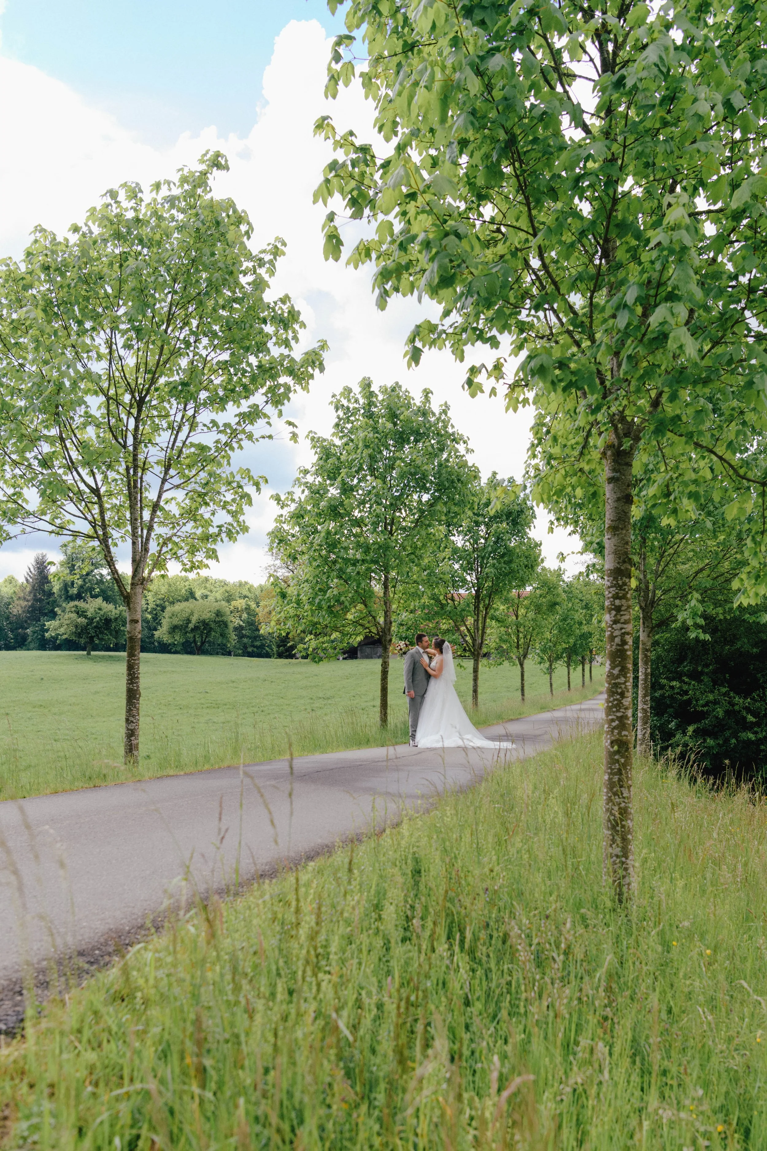 Ein Brautpaar steht auf einem Weg in einem Park mit grünen Bäumen und Wiesen im Hintergrund. Es ist ein sonniger Tag, die Szene wirkt romantisch und friedlich.