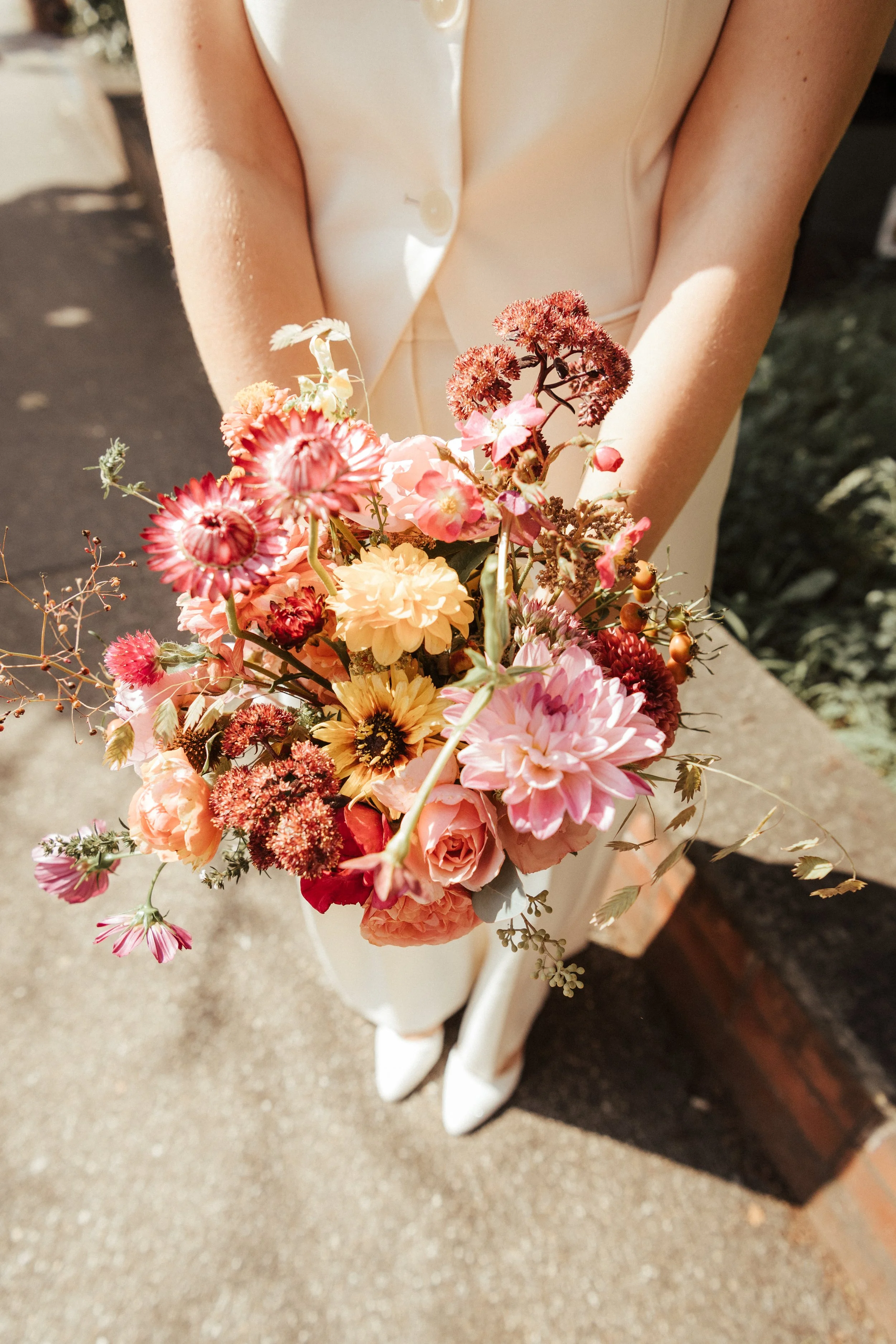 Ein Brautkleid mit weißen Schuhen, gehalten von jemandem, die einen Blumenstrauß mit bunten Blumen trägt.