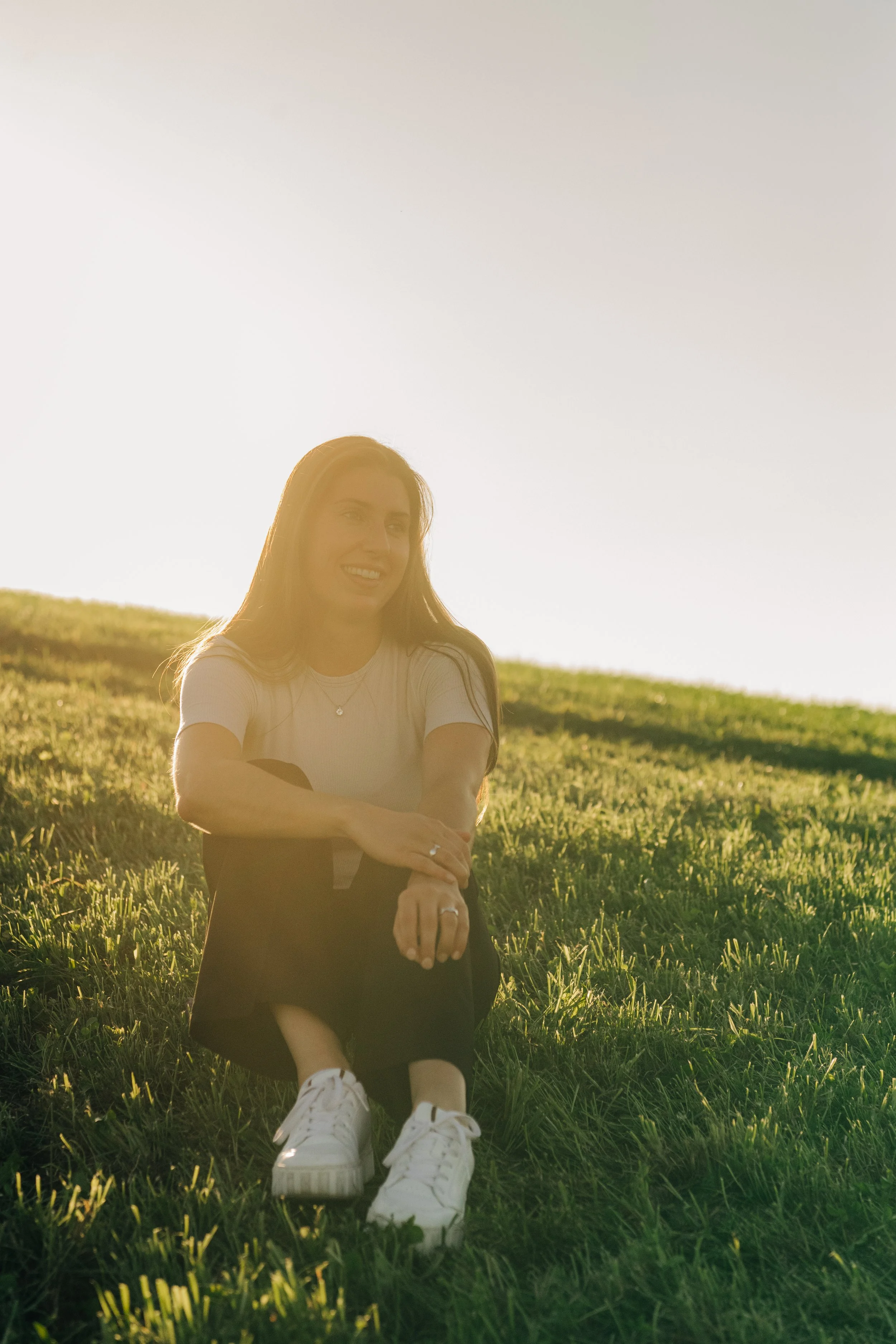 Junge Frau sitzt im Gräserfeld bei Sonnenuntergang, lächelnd, mit weißen Turnschuhen, weißem T-Shirt und dunklem Rock, in einer entspannten Pose