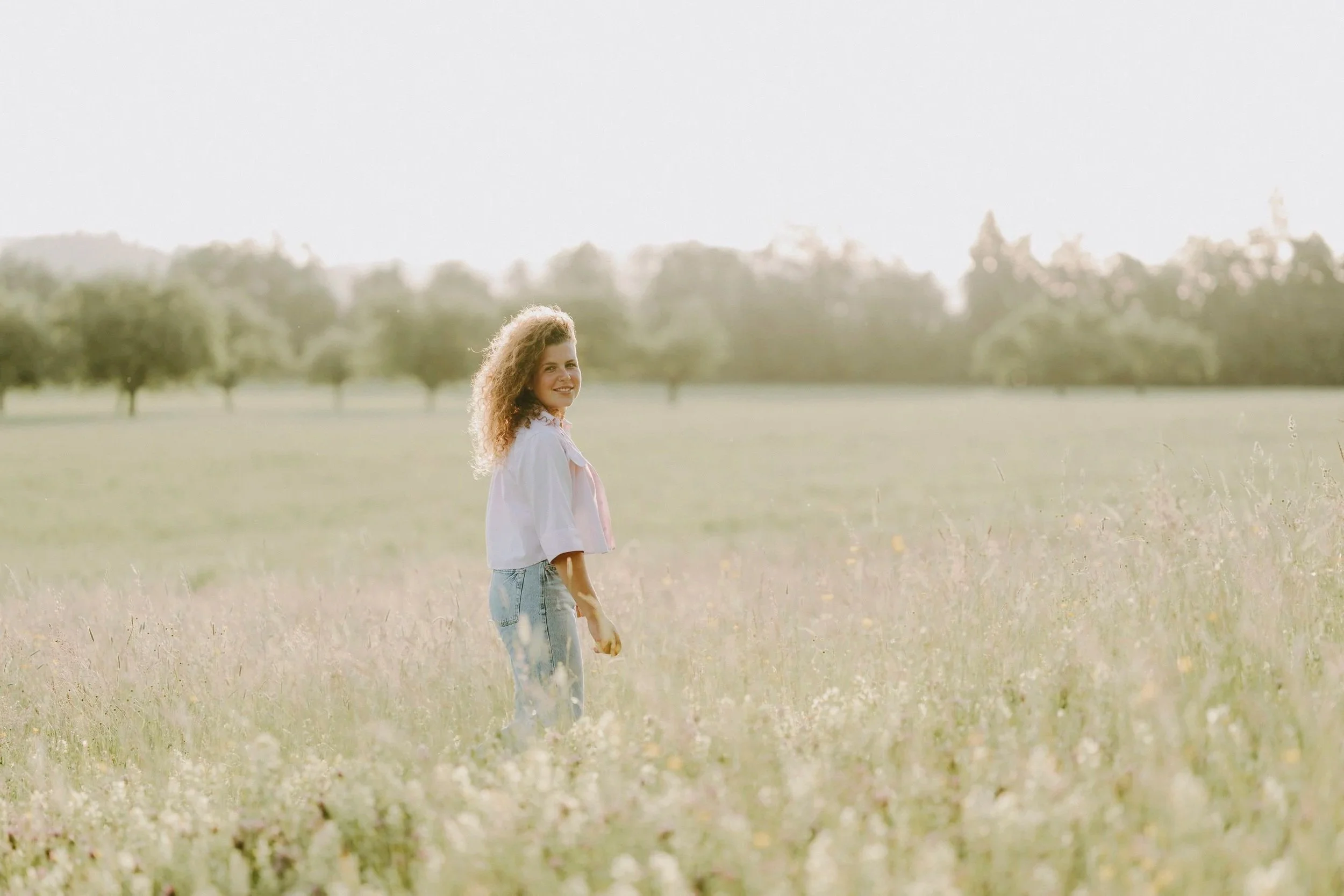 Junge Frau mit lockigem Haar, in einem hellen Hemd und Jeans, steht in einem weiten, offenen Feld mit hohen Gräsern, blicken in Richtung Kamera, bei Sonnenlicht, im Hintergrund Bäume und ein heller Himmel.