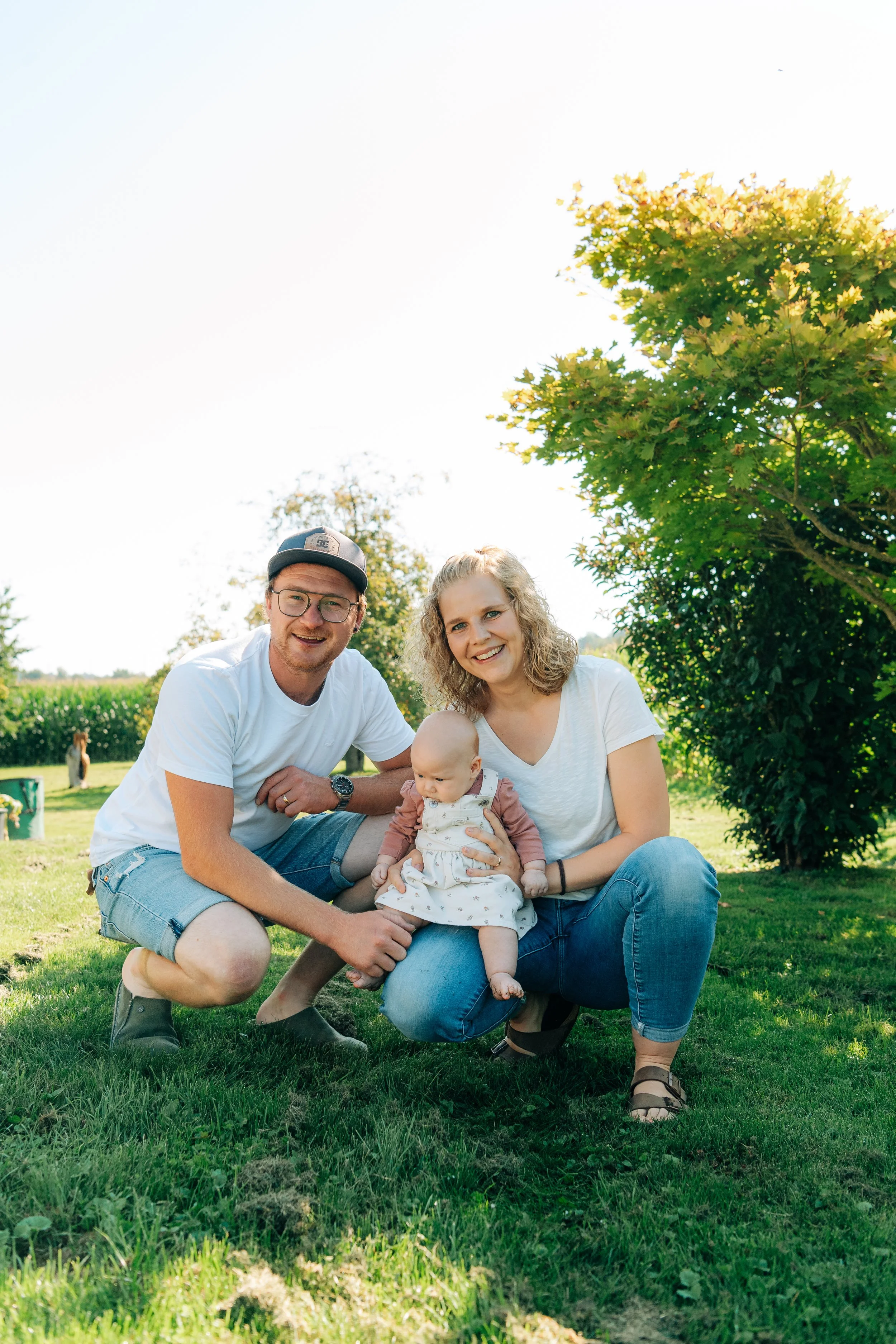 Eine glückliche Familie sitzt im Grünen in der Sonne, bestehend aus einem Mann, einer Frau und einem Baby, umgeben von Bäumen und Gras.