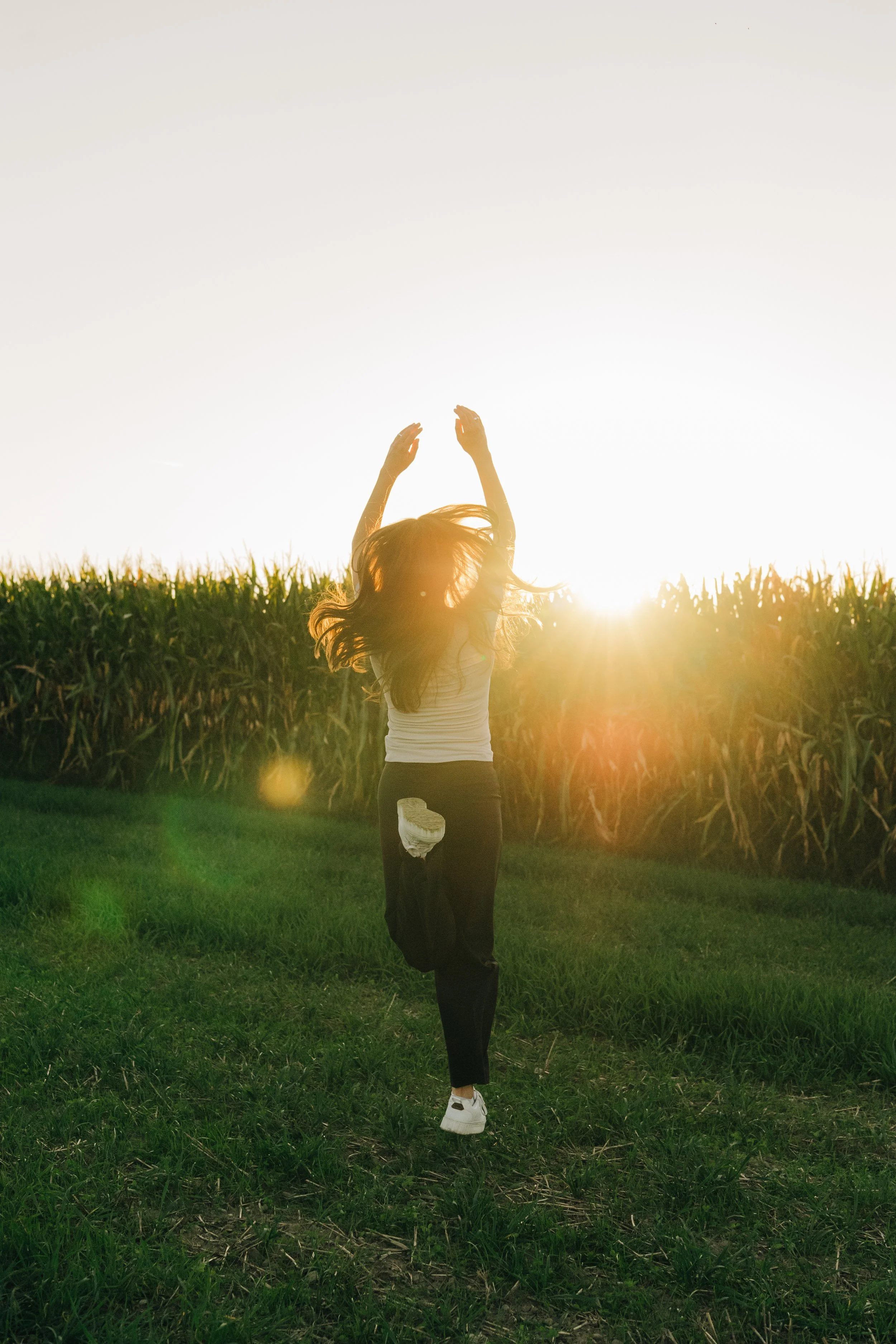 Eine Frau läuft durch ein grünes Feld in die Sonne, die im Hintergrund untergeht. Sie trägt schwarze Hosen, weiße Sportschuhe und ein weißes Top.