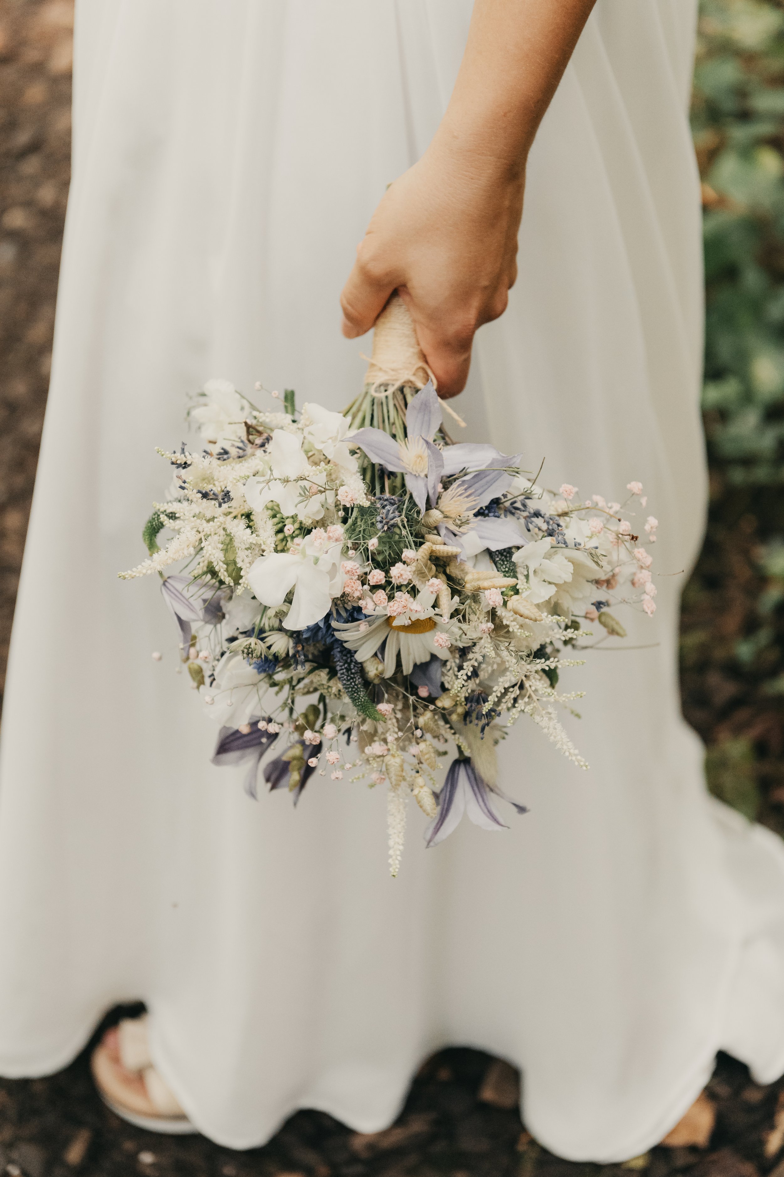 Eine Person in einem weißen Kleid hält einen Blumenstrauß mit weißen, lila, rosa und lavendelfarbenen Blumen, im Hintergrund gibt es Erde und grüne Pflanzen.
