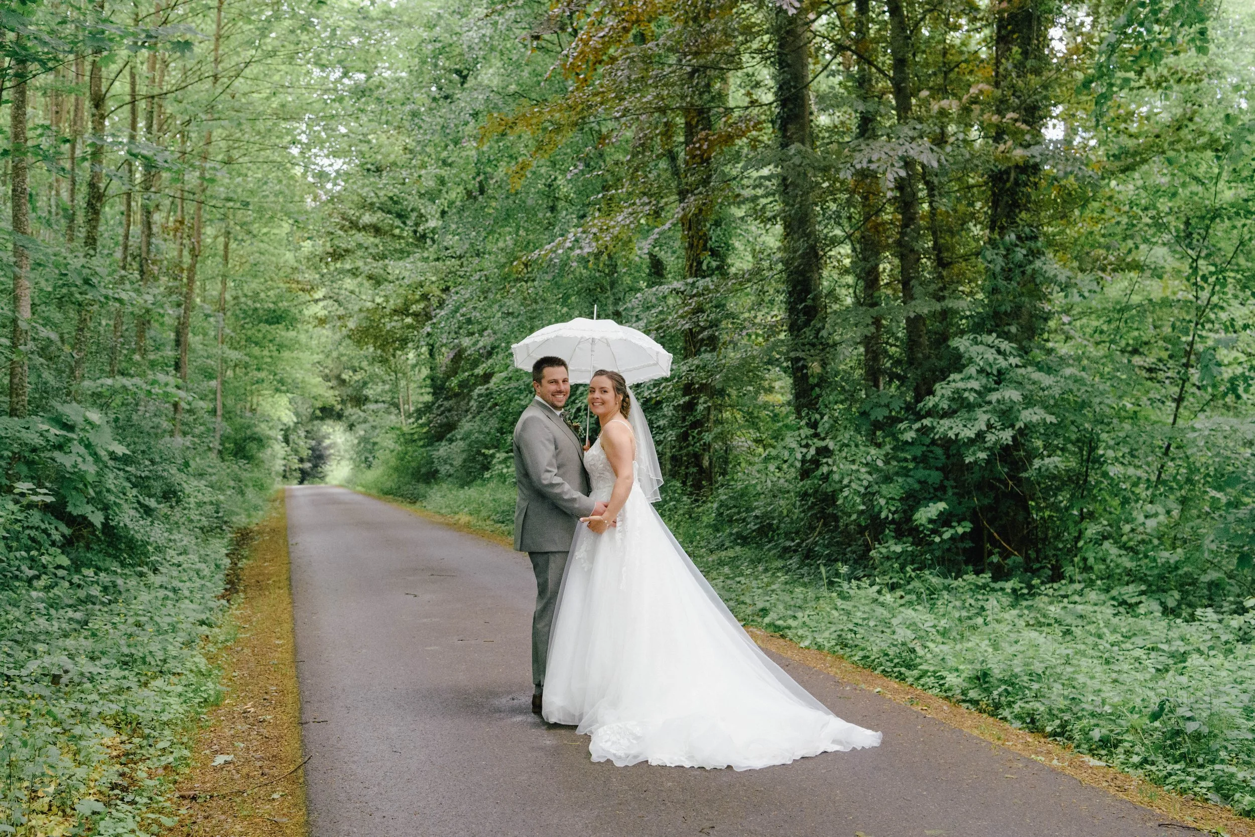 Ein Brautpaar steht auf einer schmalen, asphaltierten Straße in einem grünen Wald, der von hohen Bäumen umgeben ist. Der Bräutigam trägt einen grauen Anzug, die Braut ein langes weißes Hochzeitskleid. Der Bräutigam hält einen weißen Regenschirm über 