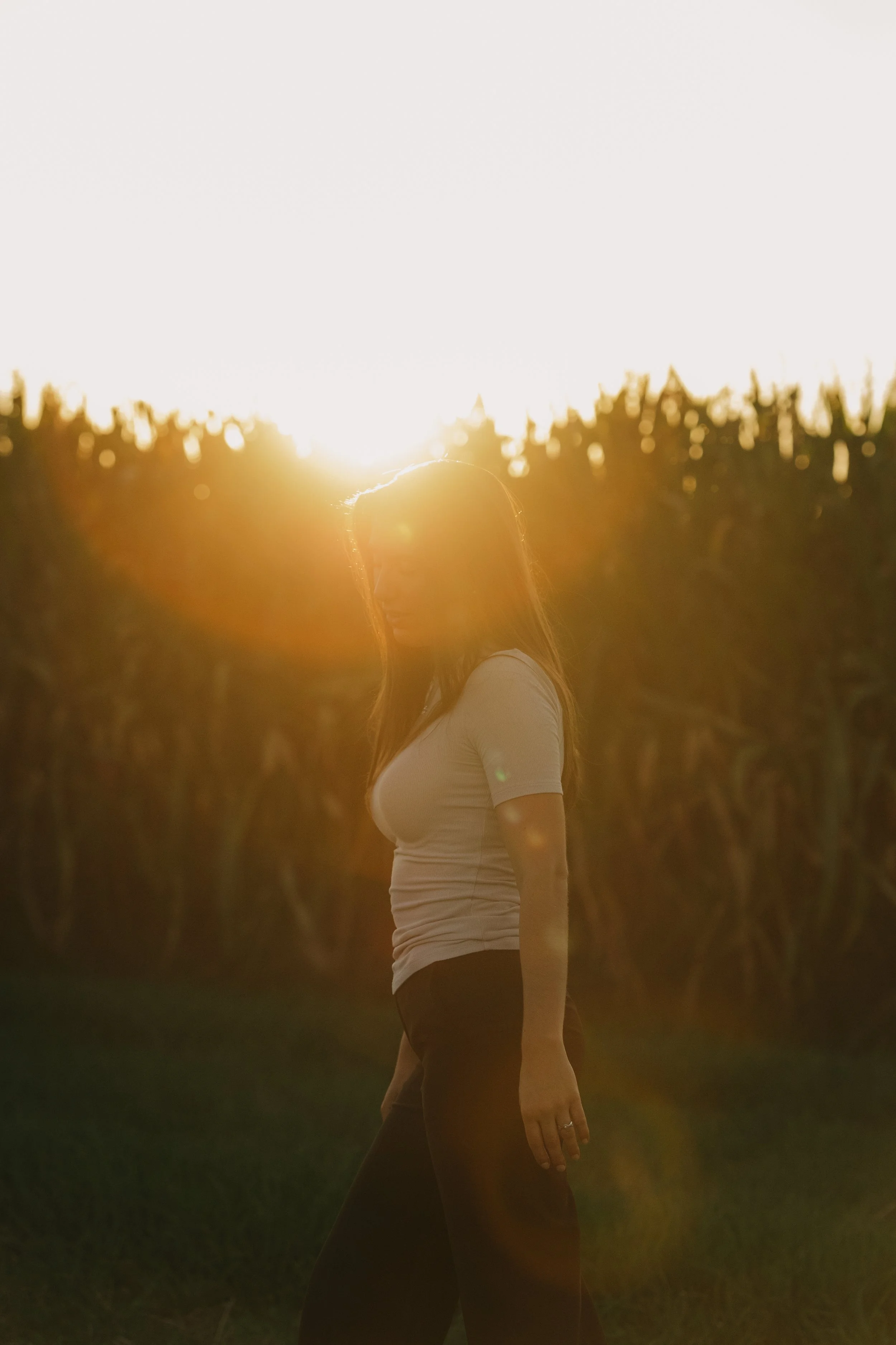 Frau steht in einem Sonnenblumenfeld bei Sonnenuntergang, Blick nach links, mit hellem Oberteil und dunklen Hosen.