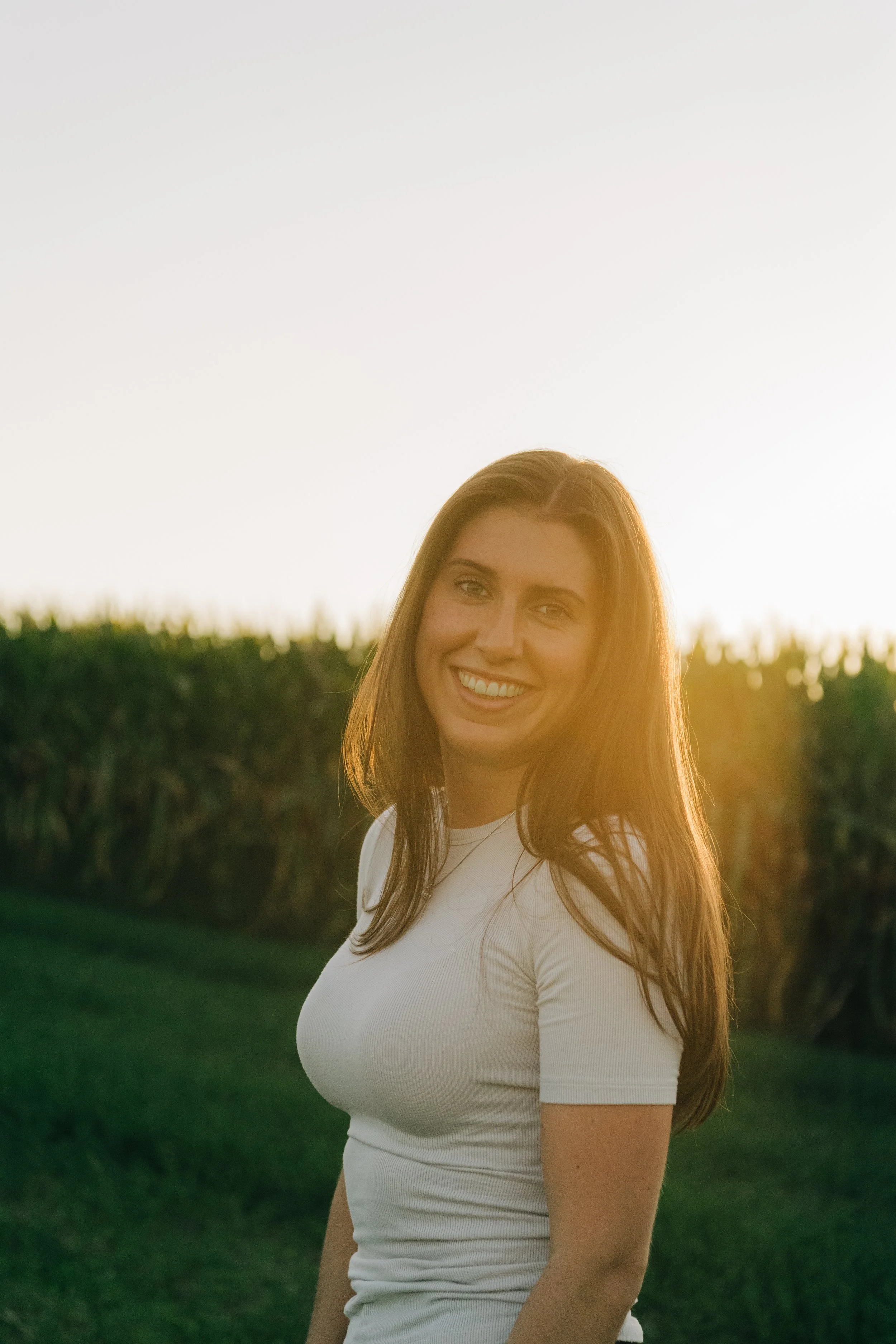 Junge Frau mit langen braunen Haaren und weißem T-Shirt steht im Freien bei Sonnenuntergang, im Hintergrund Grünflächen und Himmel.
