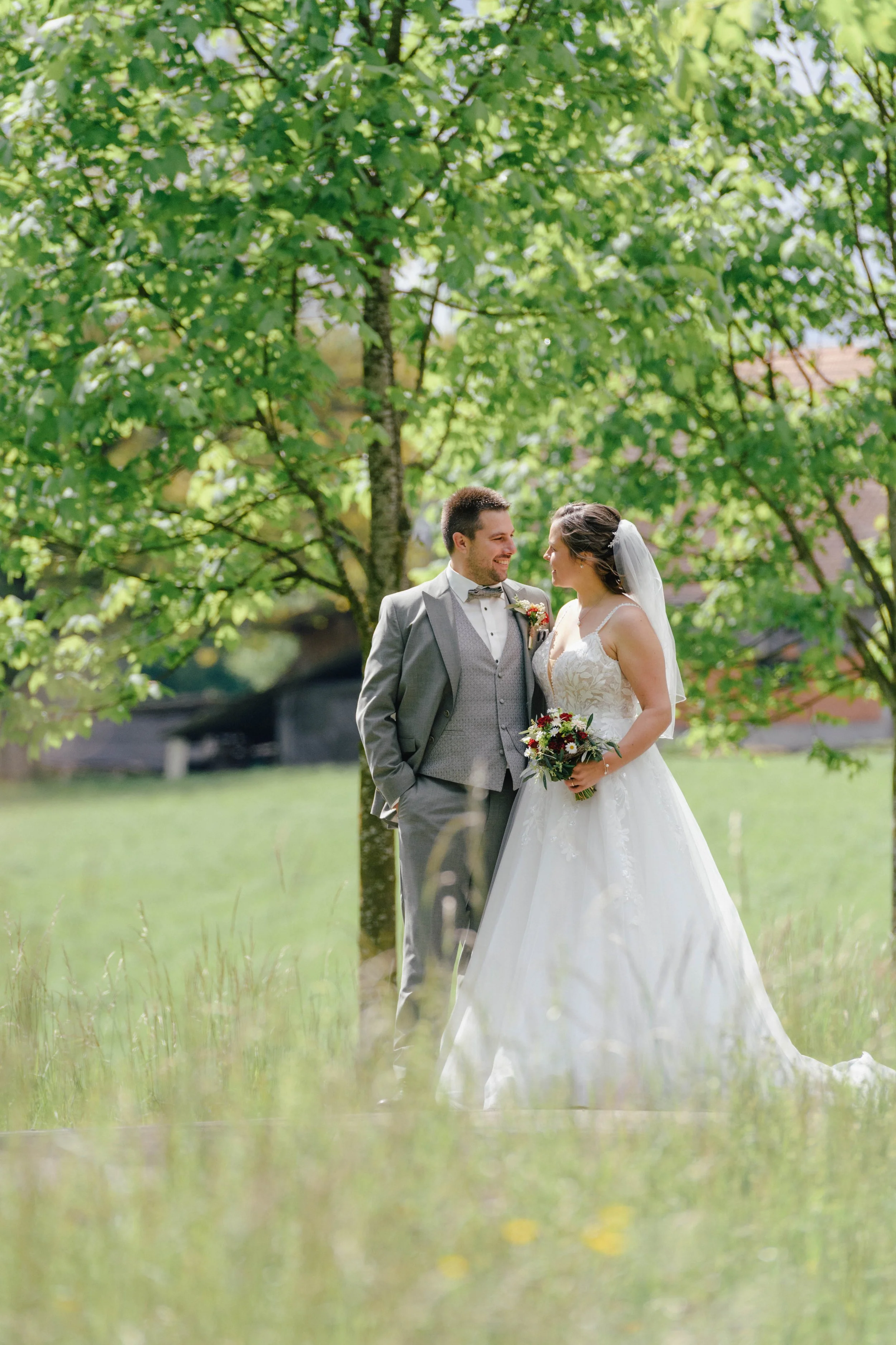 Ein glückliches Brautpaar steht unter einem Baum im Freien bei einer Hochzeit. Der Bräutigam trägt einen grauen Anzug mit Krawatte, die Braut trägt ein weißes Hochzeitskleid und einen Schleier, und sie hält einen Blumenstrauß in der Hand.