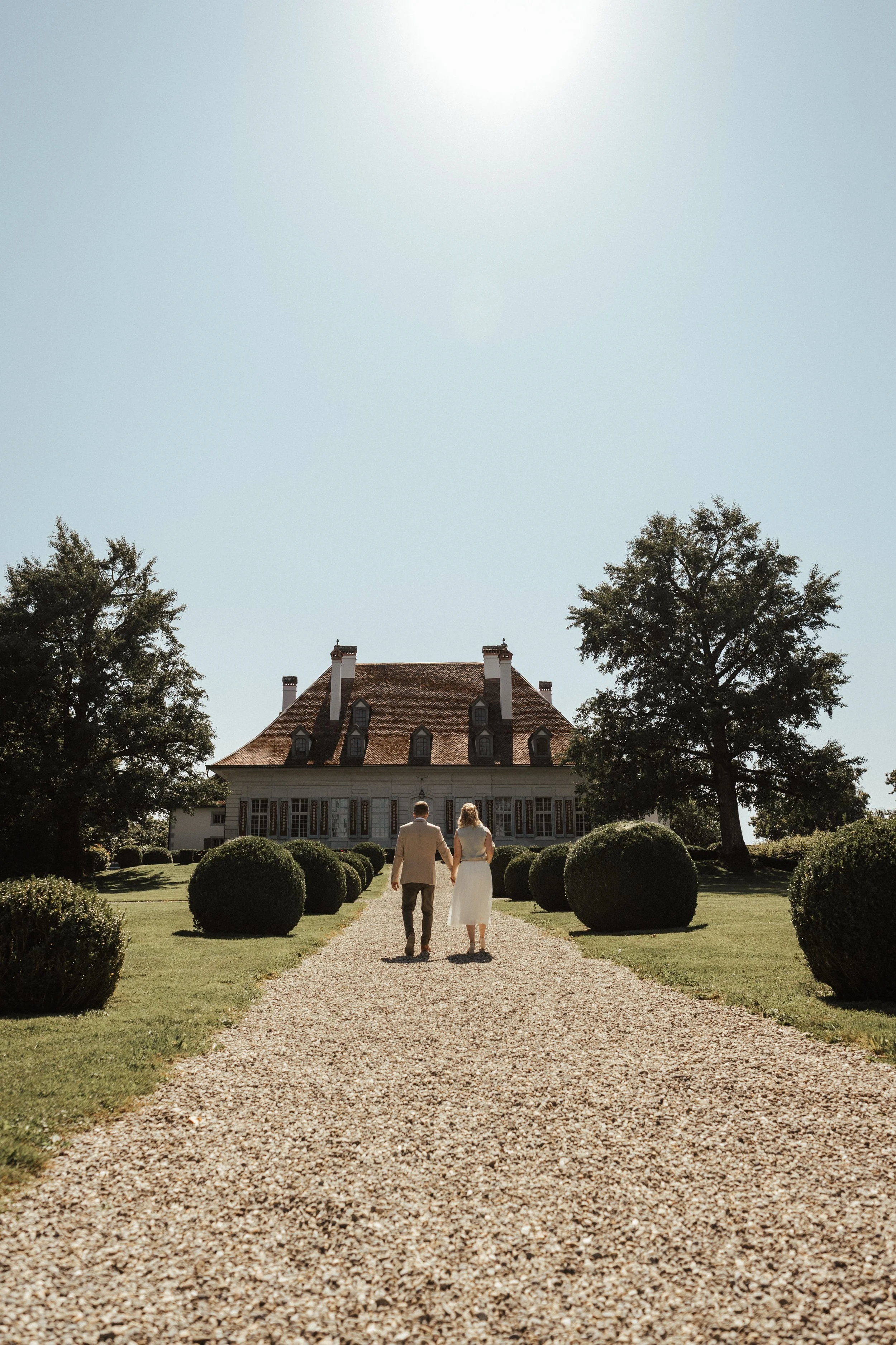 Ein verheiratetes Paar, das Hand in Hand auf einem Kiesweg zu einem großen, schönen Haus geht, umgeben von gepflegtem Rasen und runden Büschen, bei strahlendem Sonnenschein.
