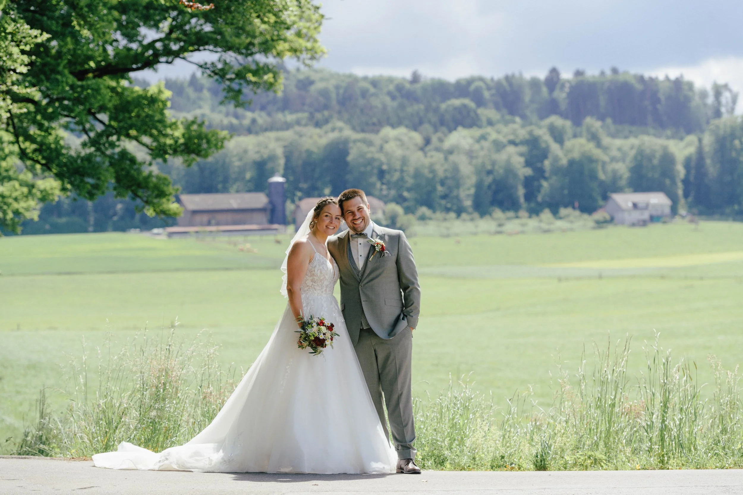 Ein glückliches Brautpaar steht im Freien vor grünen Hügellandschaften, Bäumen und einem Bauernhof im Hintergrund bei einer Hochzeit.
