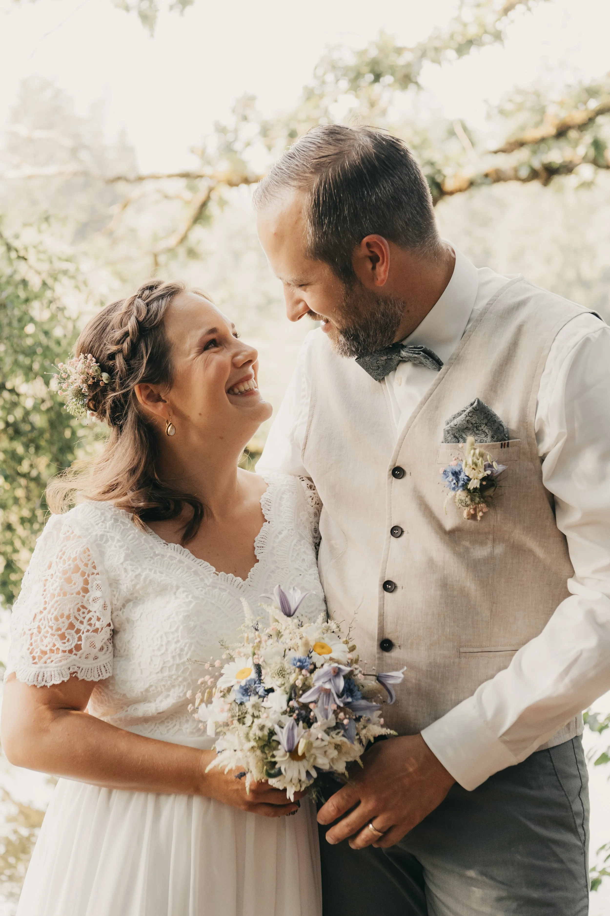 Ein glückliches Brautpaar bei ihrer Hochzeit, die Braut trägt ein weißes Kleid mit Spitze, sie hält einen Blumenstrauß, der Bräutigam trägt einen beige Weste mit Blumenanstecker und eine Fliege, sie lächeln einander an, im Hintergrund sind Bäume und 