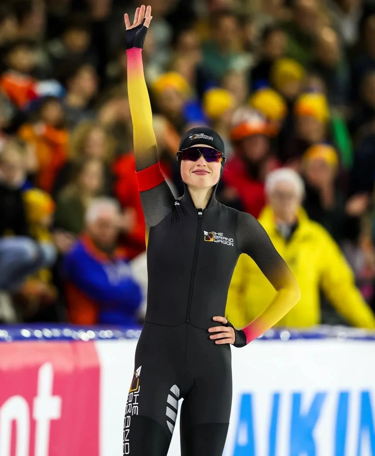 De vrouw in een zwart sportoutfit met gele en roze mouwen staat met één hand in de zij en zwaait met de andere hand omhoog. Ze draagt een zwarte helm, zonnebril, en een glijdende rits op haar sportoutfit, met een grote glimlach op haar gezicht terwijl ze in een schaatsbaan staat. Er staat een grote groep mensen achter haar, gekleed in kleurrijke ski- of sportkleding.