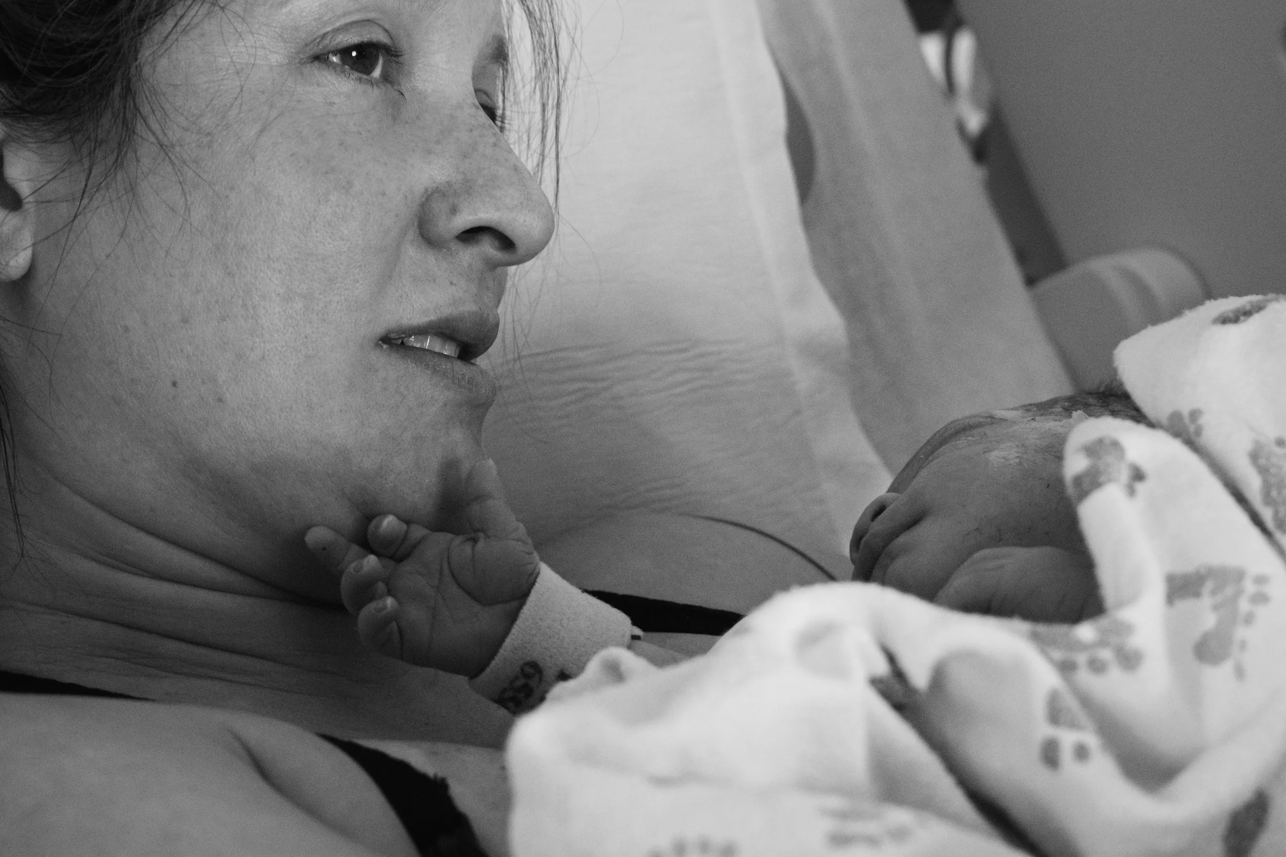Close-up of a woman with freckles looking intently at a baby, who is holding a piece of food and wearing a hospital bracelet, in black and white.