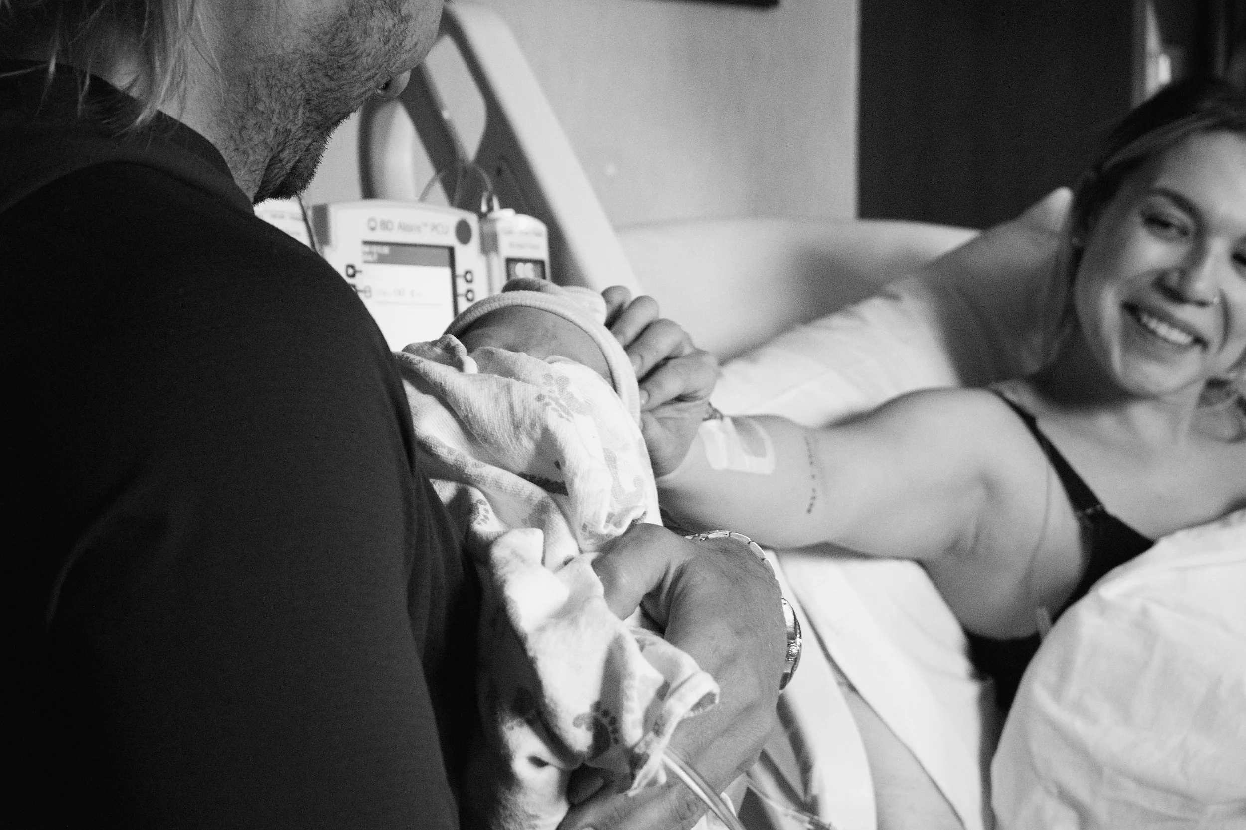A woman lying in a hospital bed smiling while a man with a beard and long hair holds her arm, possibly during a medical procedure or checkup, in a clinical setting.