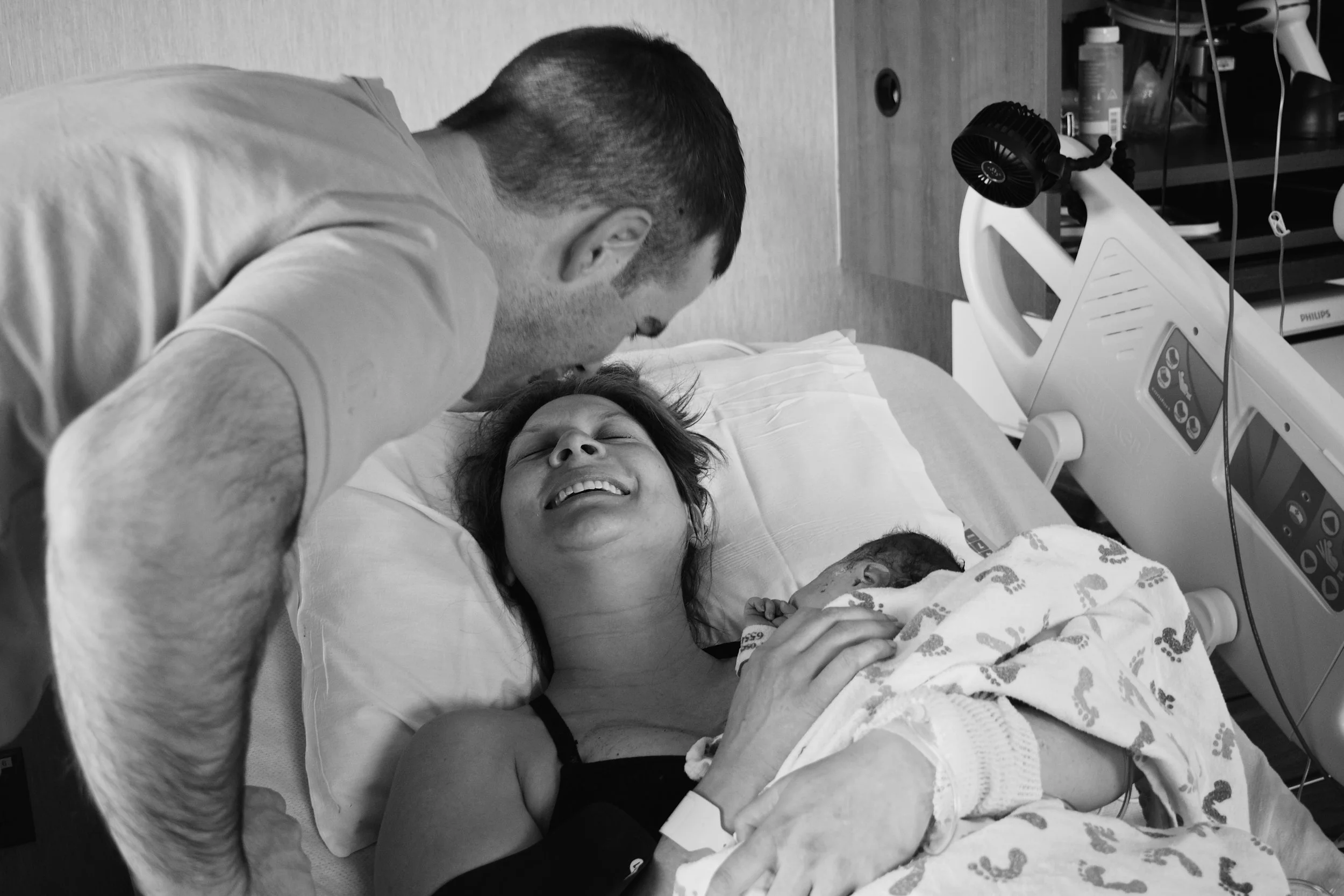 A woman lying in a hospital bed, smiling and holding her newborn baby, as a man leans over her in a hospital room.