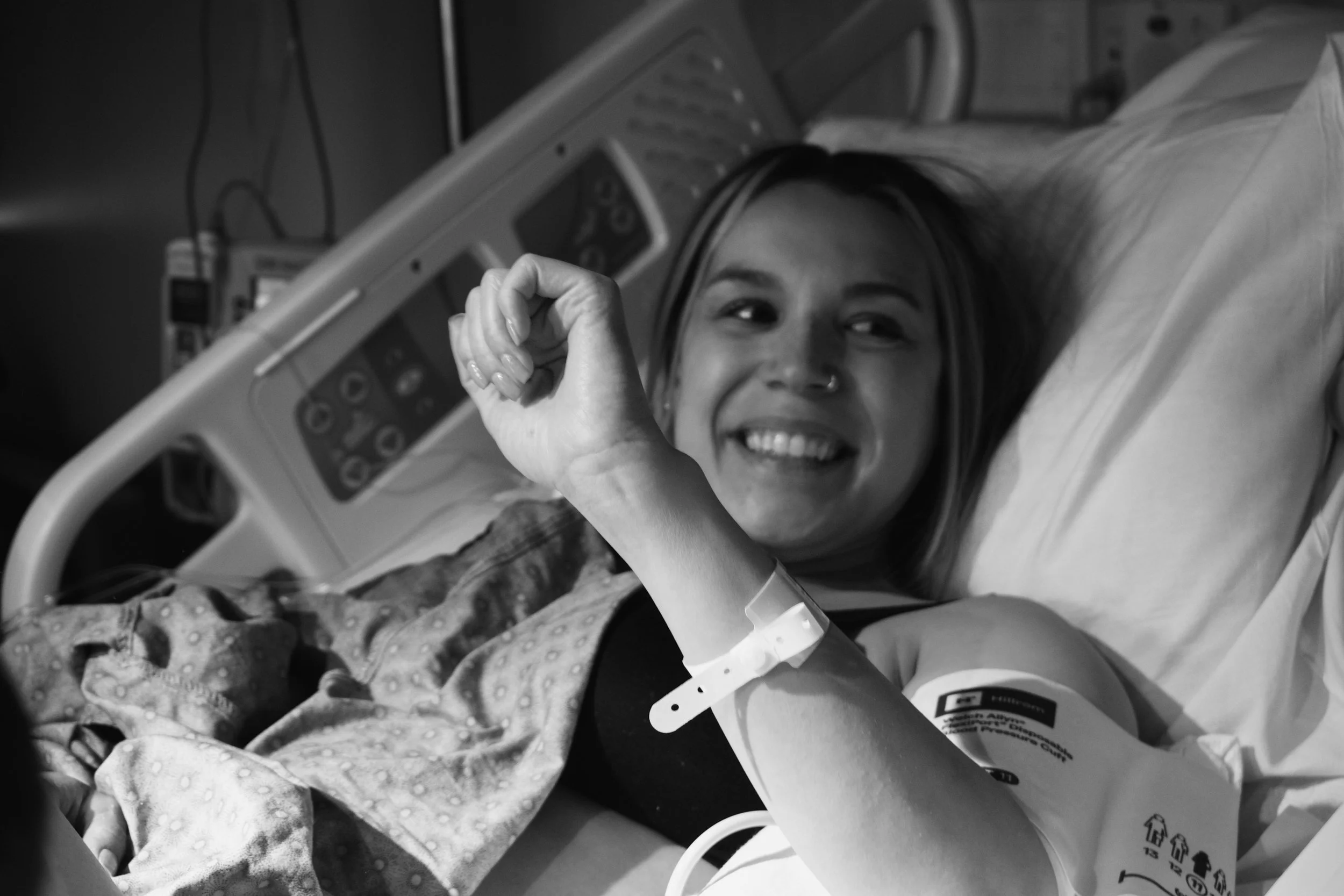 A woman lying in a hospital bed smiling and making a fist with her right hand, wearing a hospital wristband, with medical equipment visible around her.