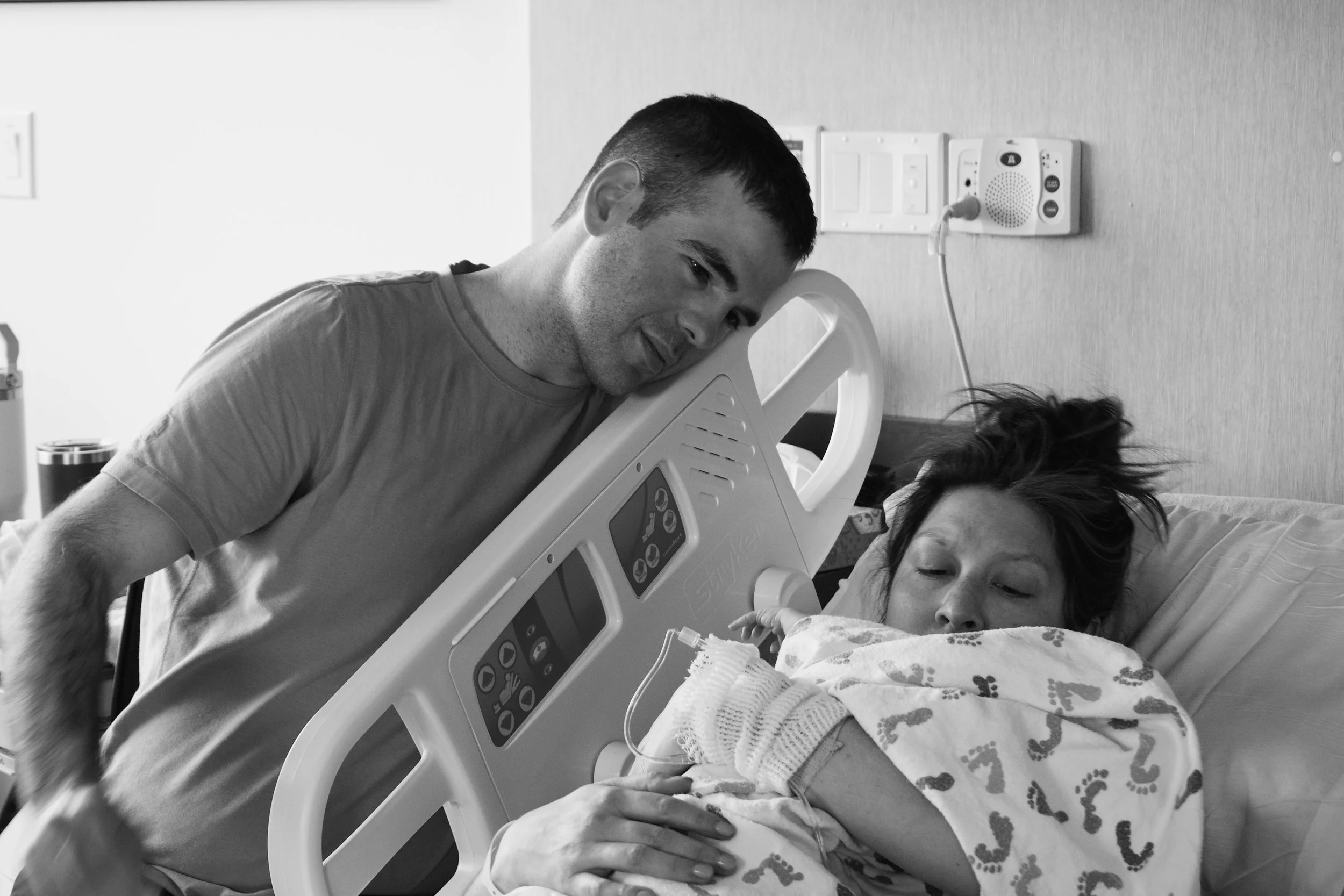 Man leaning on hospital bed next to a woman lying in bed, holding her hand, in a hospital room.