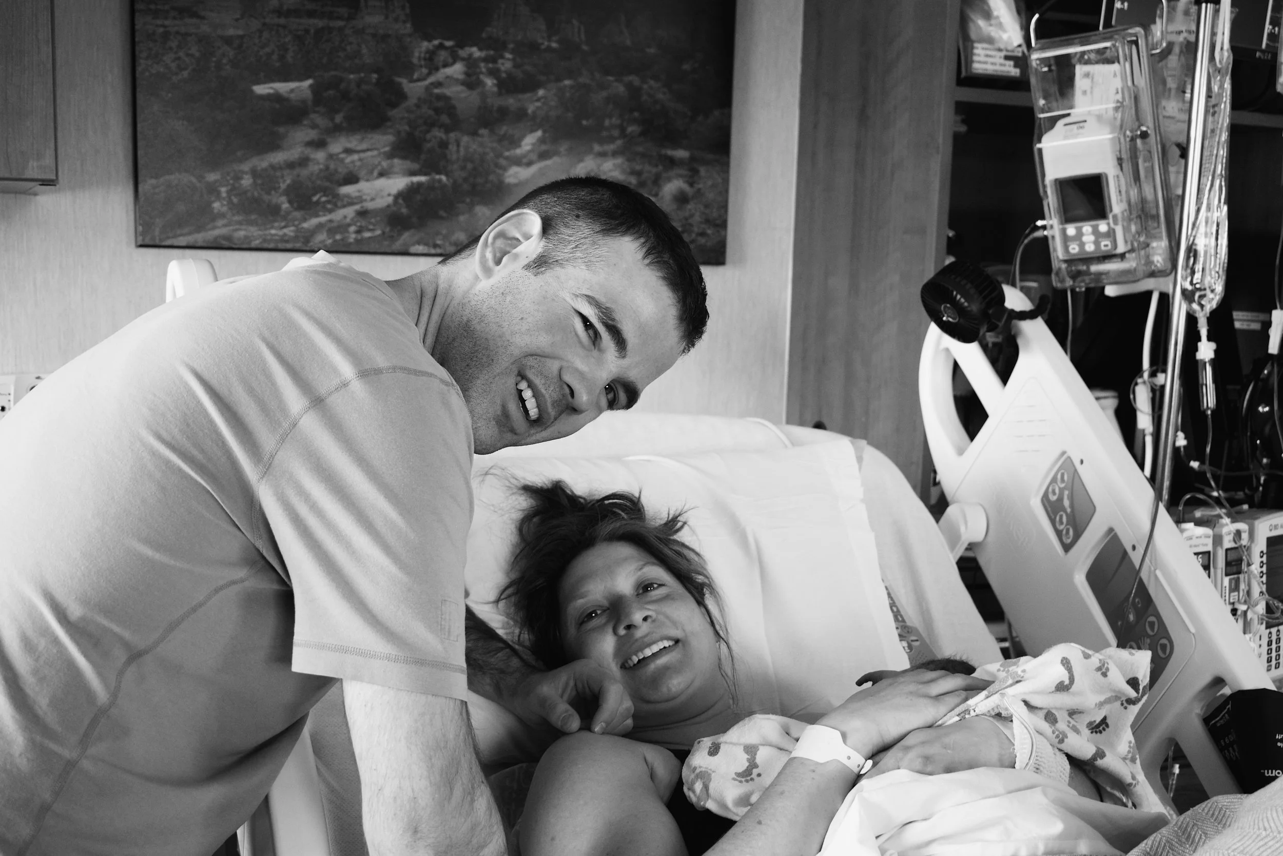 A man and woman in a hospital room, with the woman lying in a bed with medical equipment; the man is leaning over and smiling at her.