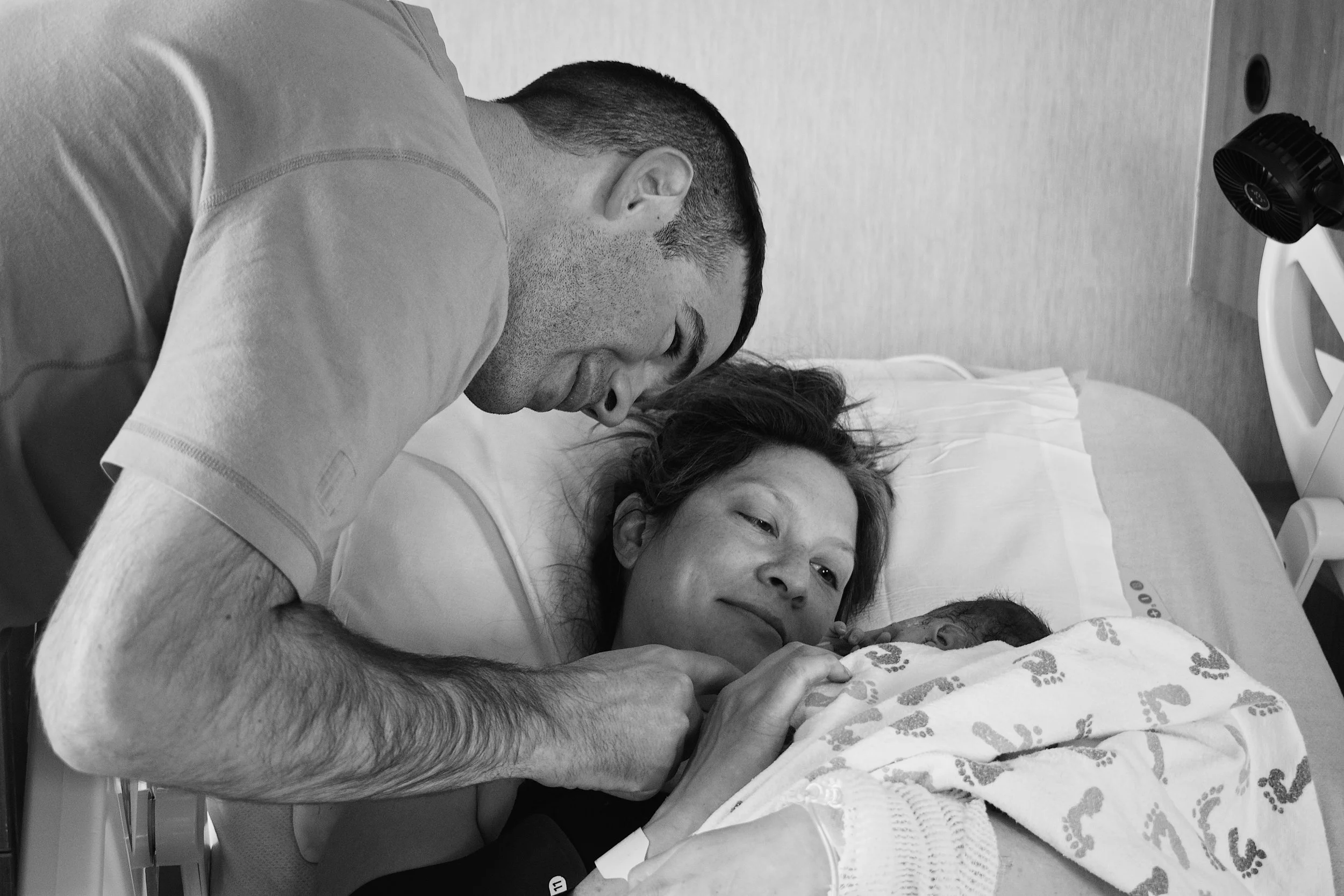 A man with short dark hair leaning over a woman lying in a hospital bed, holding her hand as she gazes at him. The woman has dark hair and is partially covered with a patterned blanket. Medical monitors are visible in the background.