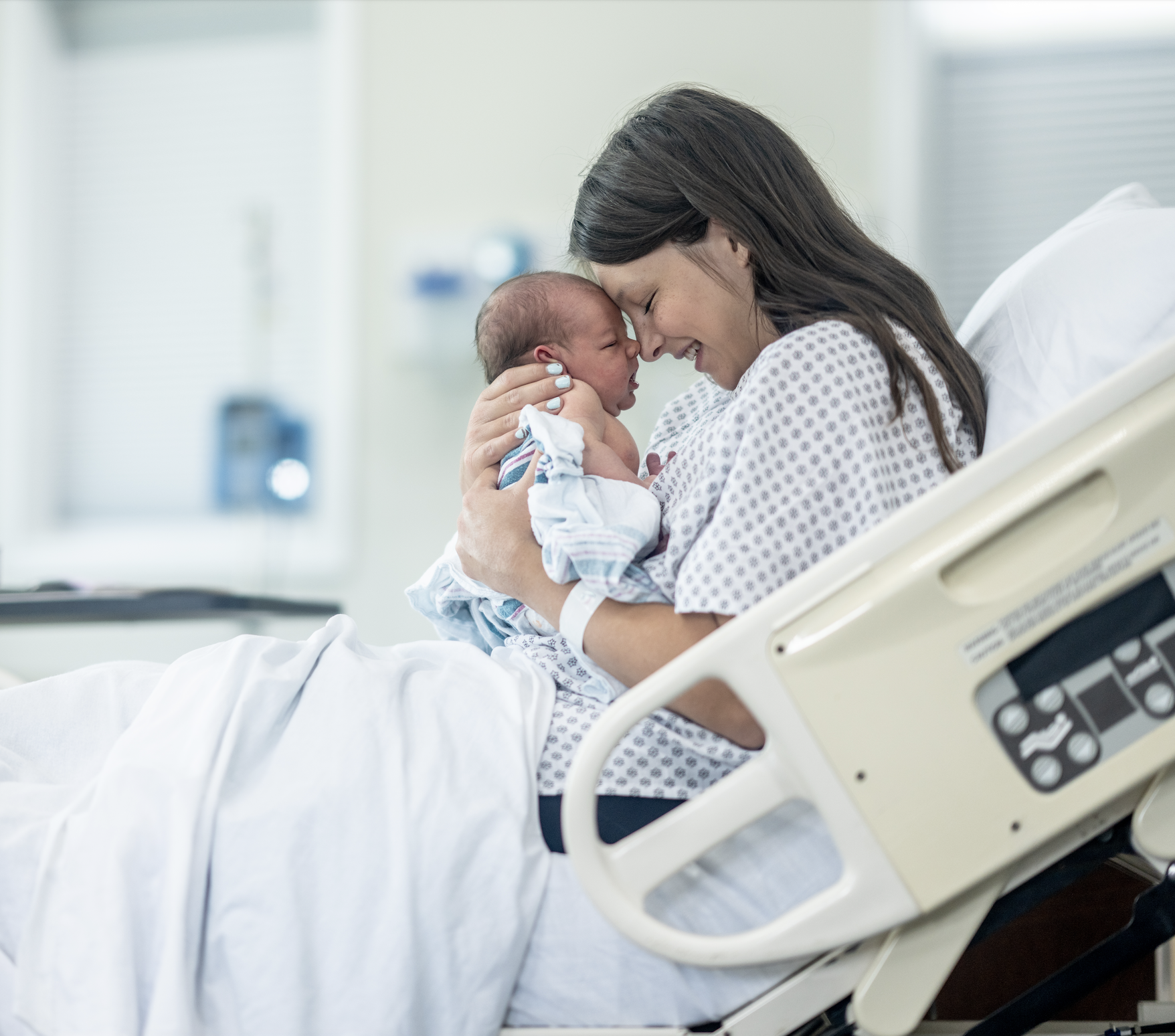 A woman in a hospital gown smiling and holding a newborn baby in a hospital bed.