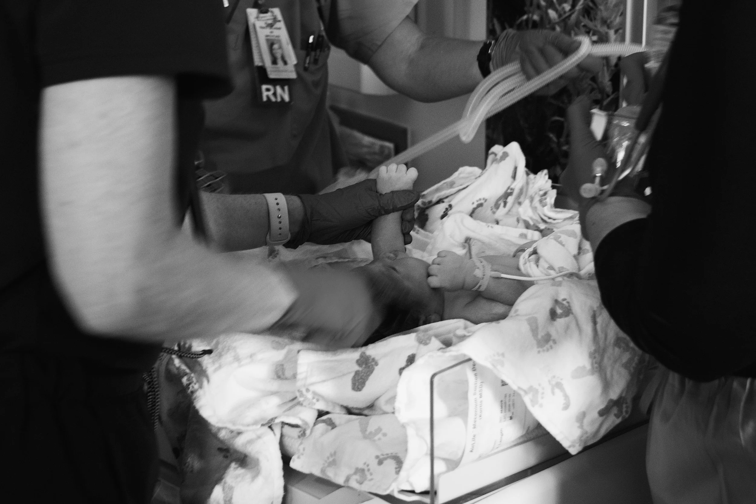 A healthcare professional, wearing gloves and a medical badge, holding a newborn baby in a hospital setting, with hospital equipment and a hospital bed in the background.
