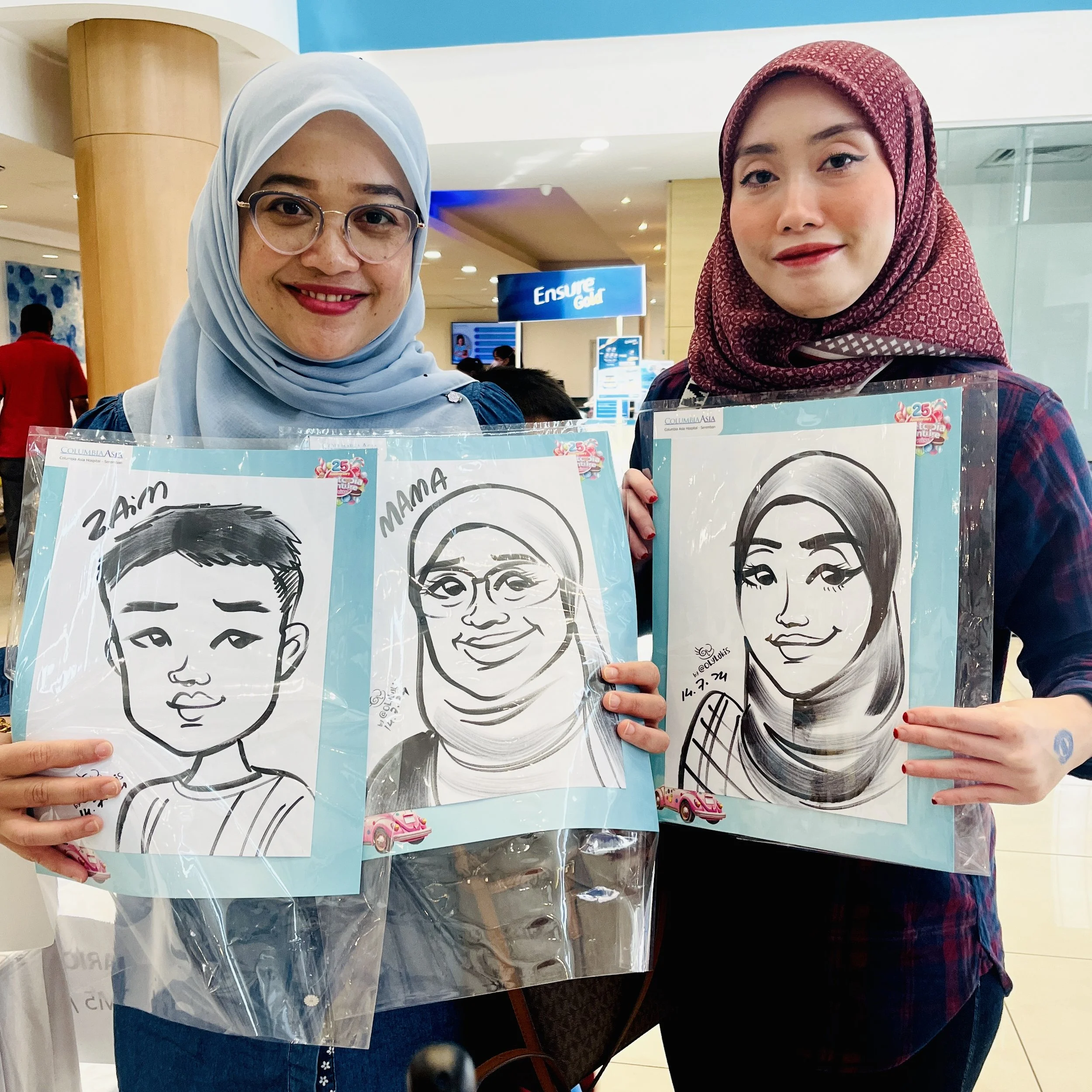 Two women with headscarves holding caricature drawings of themselves at an indoor event.