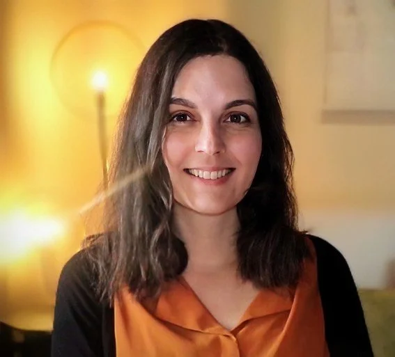 A woman smiling, with shoulder-length dark hair, wearing a black and orange top, indoors with warm lighting.