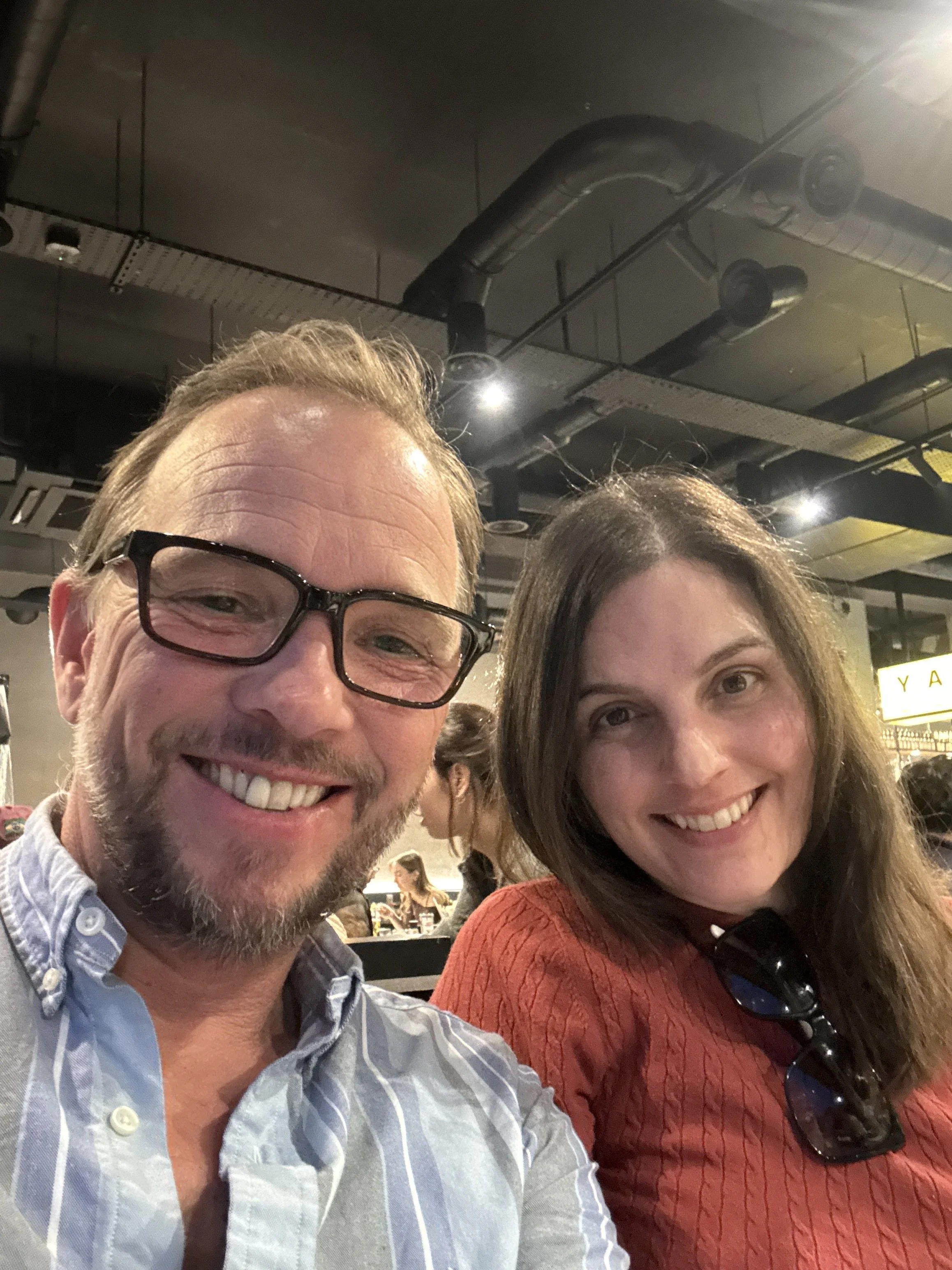 Close-up of a smiling man with glasses and beard, and a smiling woman with long hair, sunglasses hanging on her shirt, in a restaurant or cafe.
