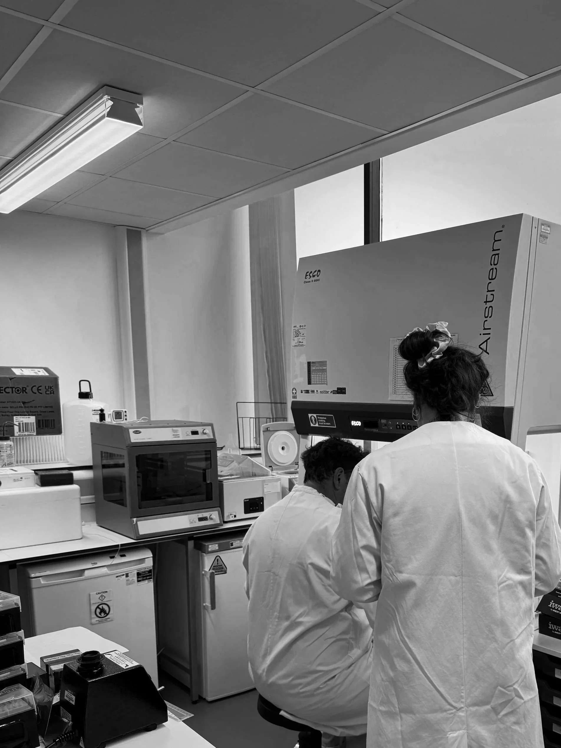 Laboratory with two scientists in white coats working at a bench with medical and electronic equipment, including monitors and large devices labeled ESCO and Airstream, in a well-lit room with a window.