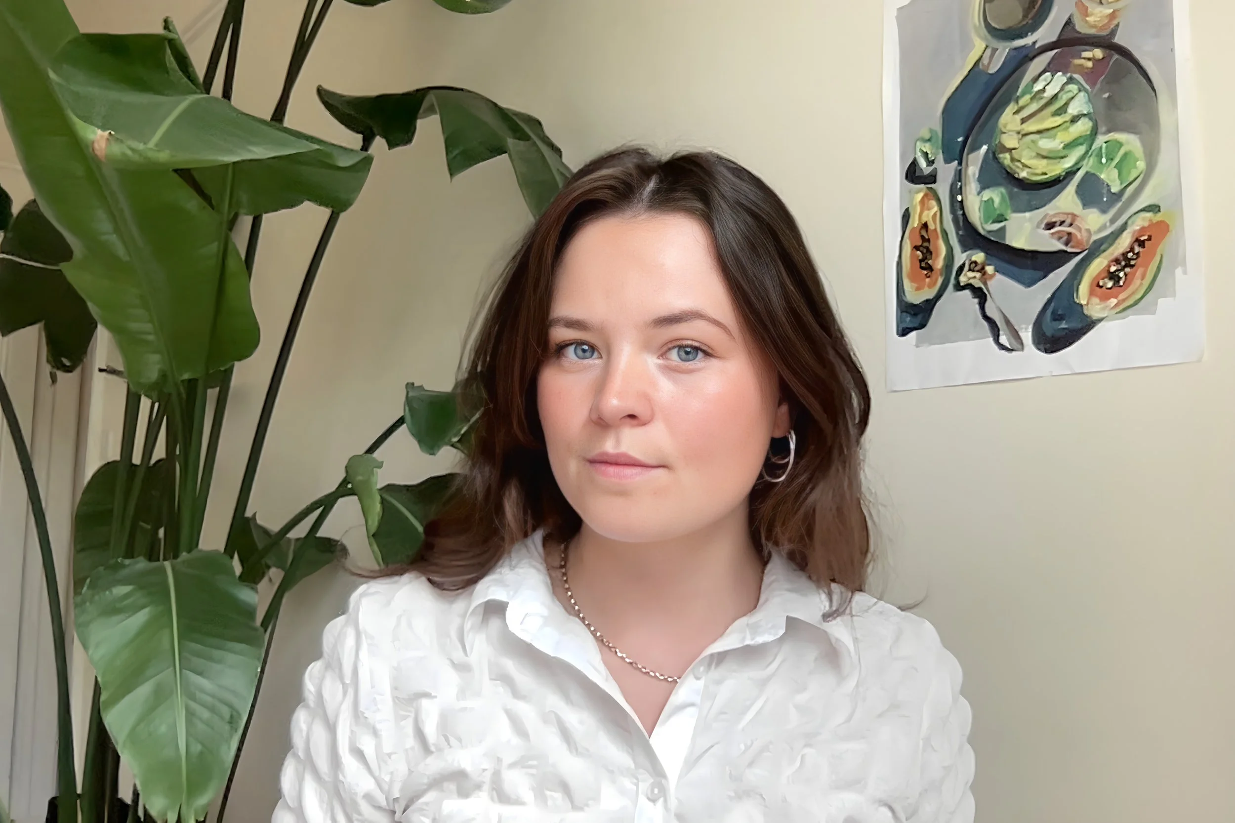 Close-up of a young woman with light skin, brown hair, and blue eyes, wearing a white blouse and silver jewelry, standing indoors next to a large green plant and a colorful abstract painting on the wall.